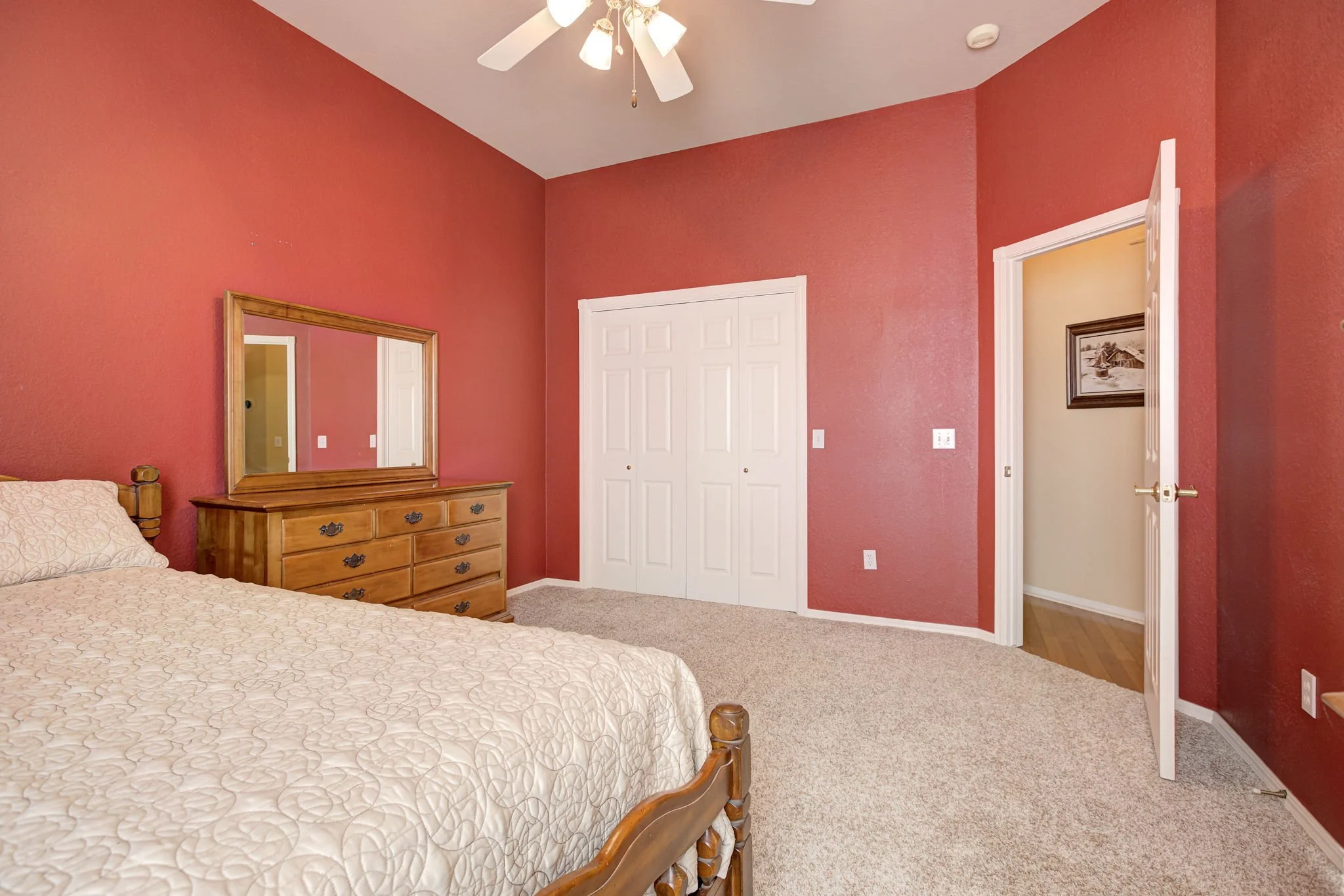 A bedroom with red painted walls, a wooden bed with a patterned bedspread, a wooden dresser with a large mirror, a white double closet door, a ceiling fan with lights, and a framed picture on the wall visible through an open door.