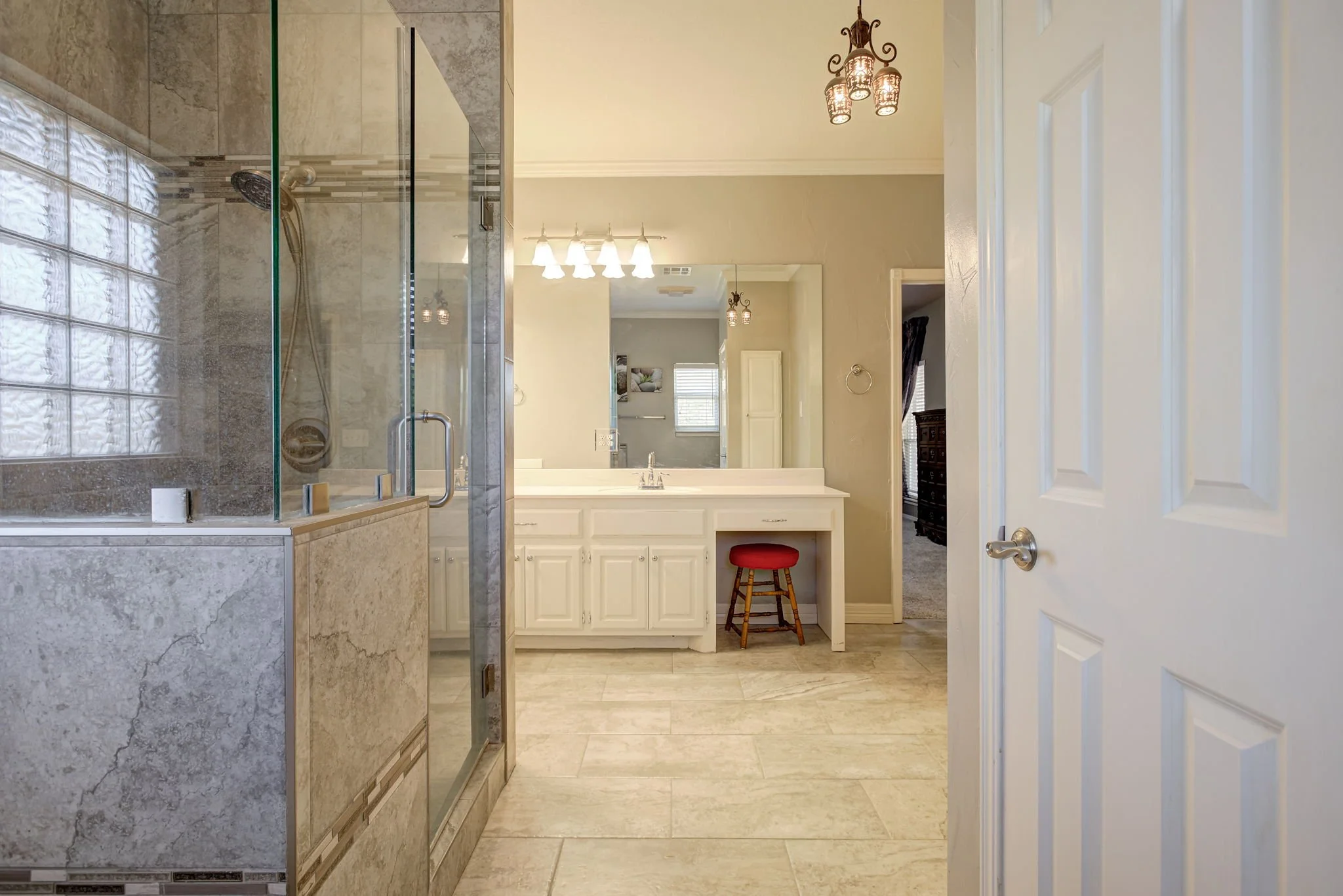 View of a bathroom with a shower on the left, a large mirror, a vanity with a sink, and a small red stool. The ceiling has three hanging light fixtures, and there is a door on the right side of the image.