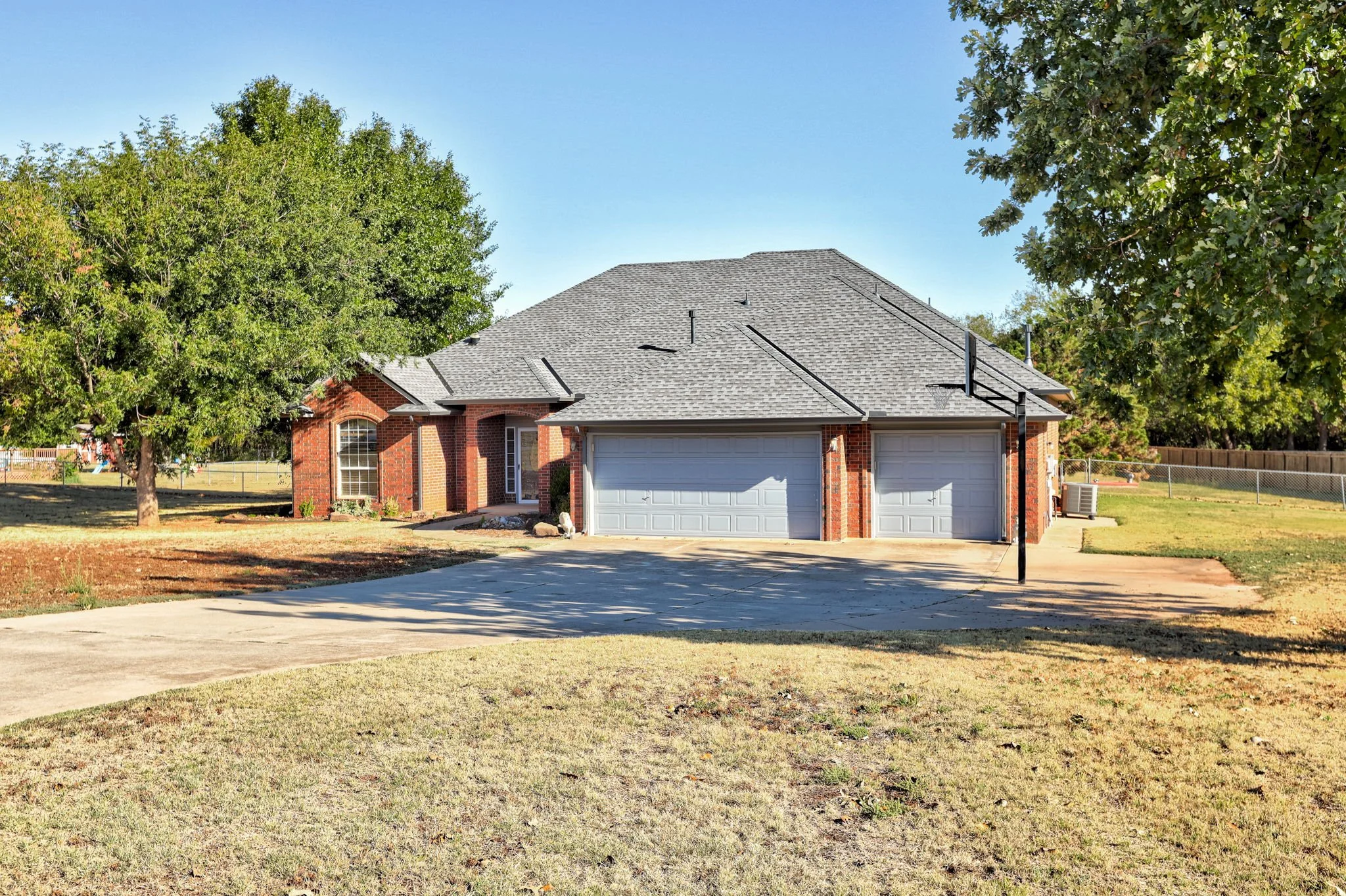 A brick house with a gray shingle roof, two-car garage, surrounded by large green trees, with a fenced backyard and a clear blue sky.