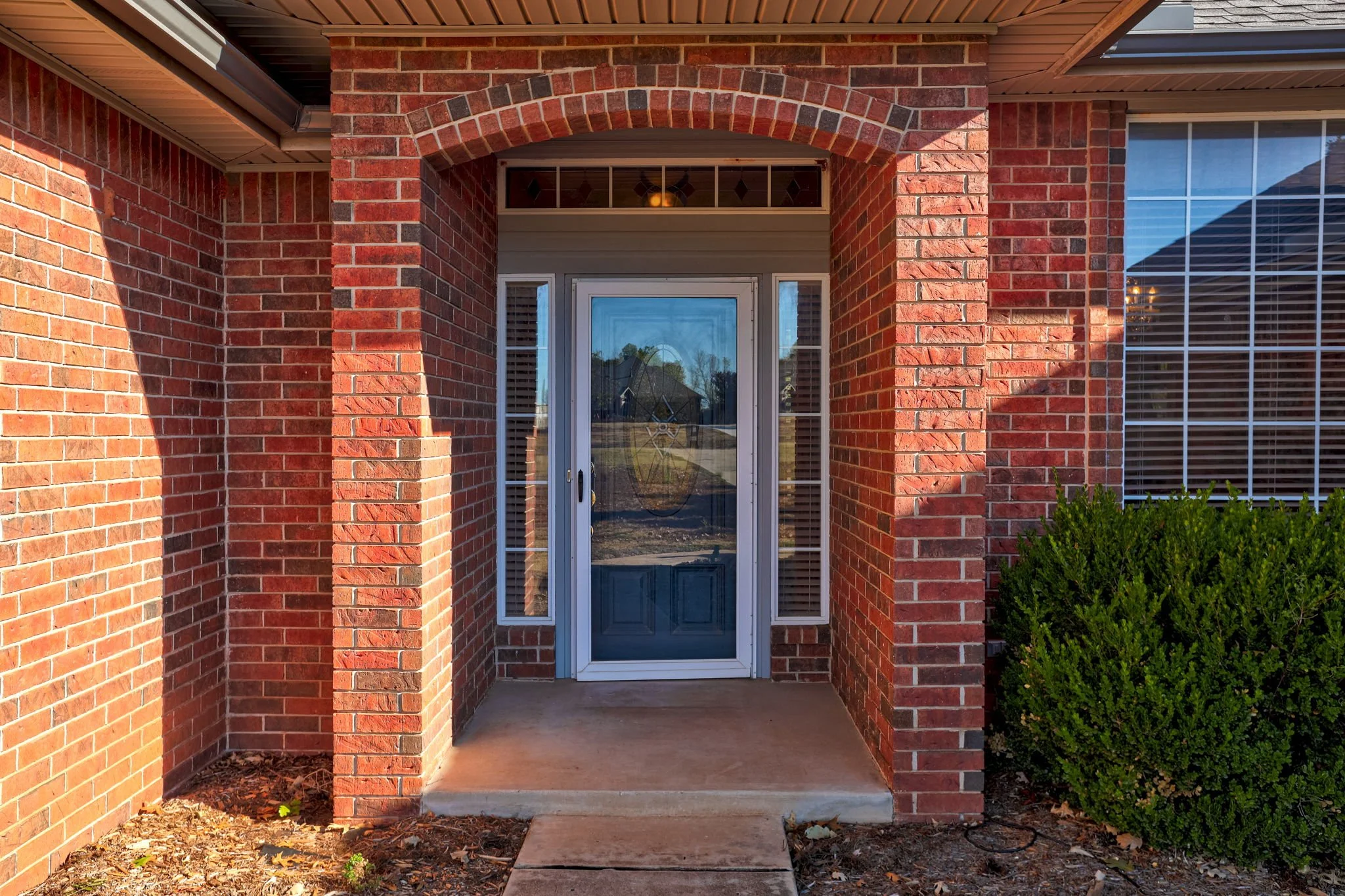 Front entrance of a brick house with a covered porch, front door with a glass storm door, side window blinds, and a bush on the right.