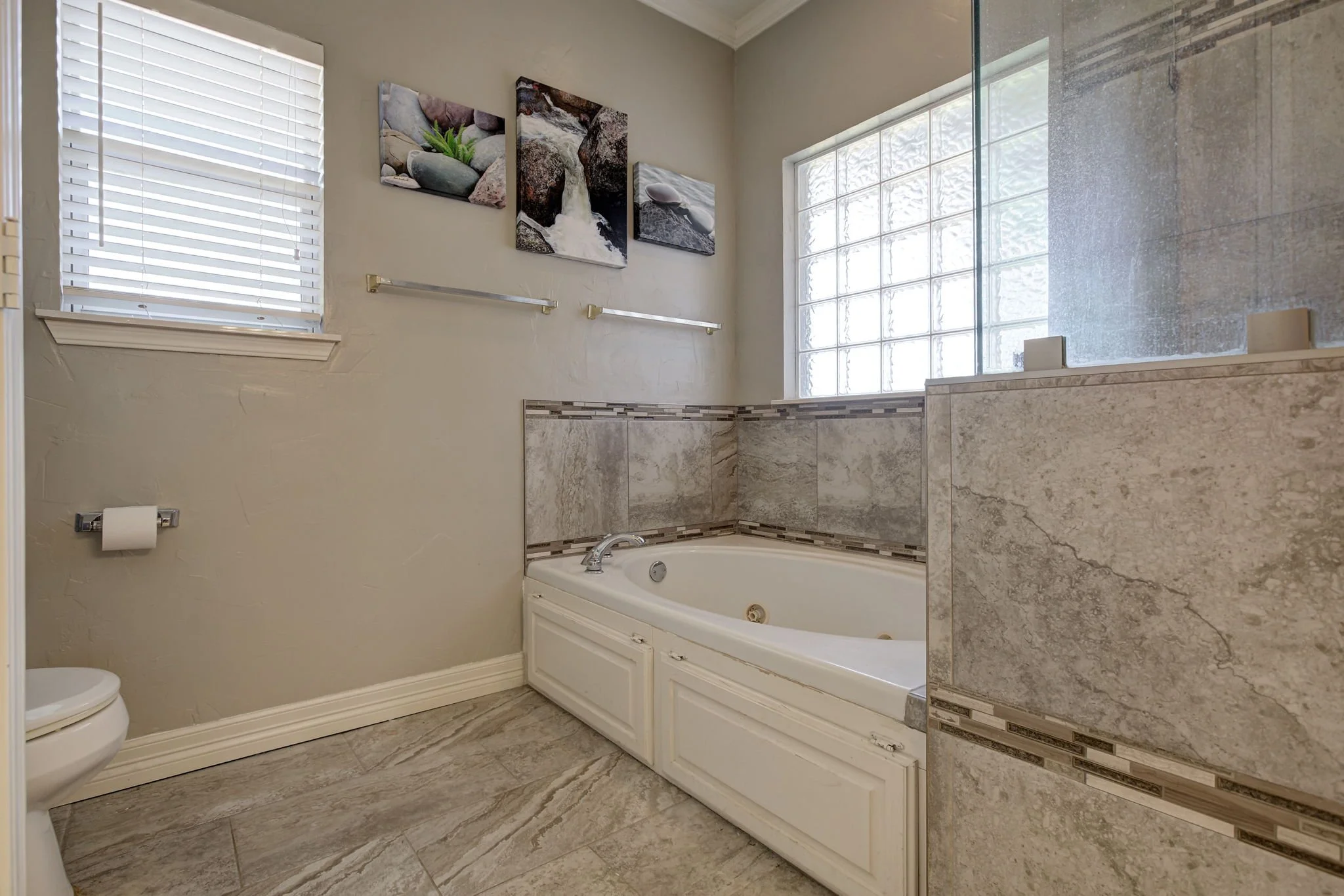Bathroom with a bathtub, tiled walls, a large glass block window, and window blinds. There are three canvas prints of rocks and water on the wall and two grab bars above the tub.