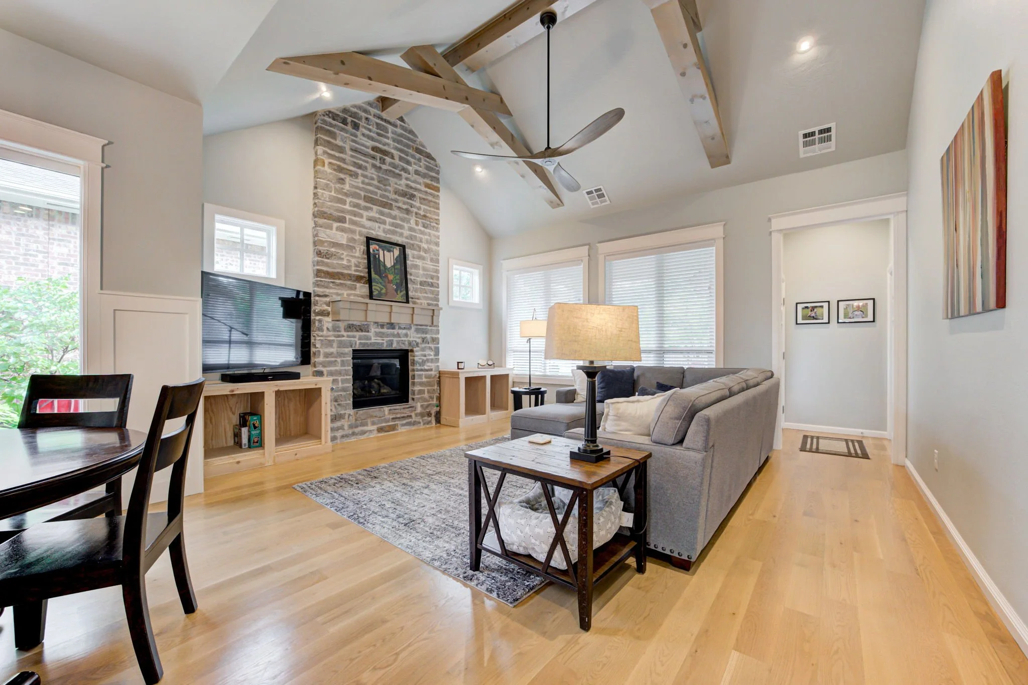 Living room with light hardwood floors, a gray sectional sofa, a wooden side table with a lamp, a stone fireplace, a flat-screen TV, and multiple windows with white blinds.