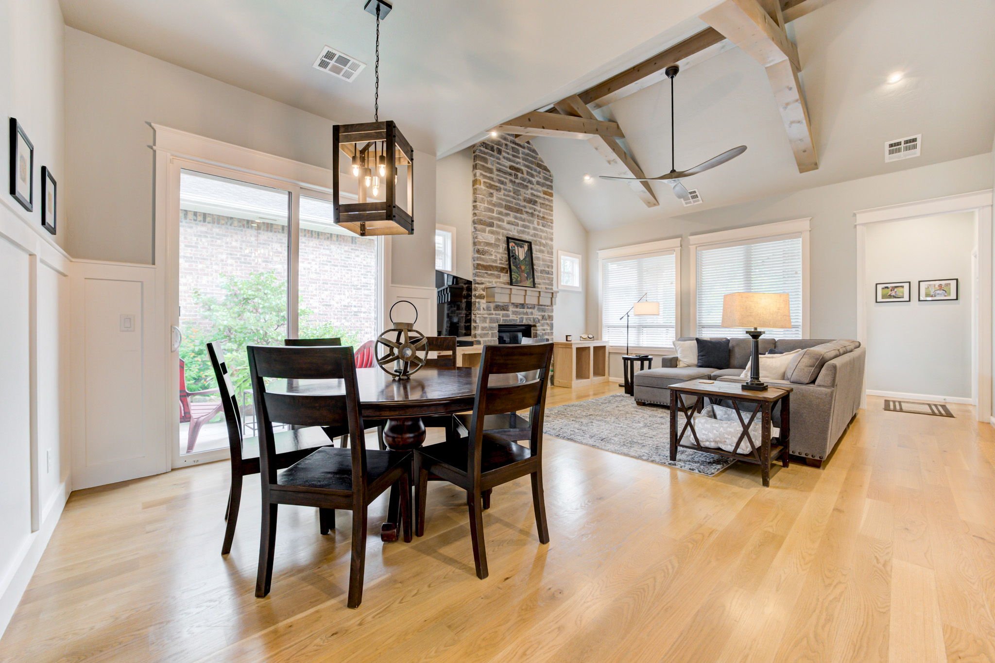 Living room with light wood flooring, a stone fireplace, and a vaulted ceiling with exposed wooden beams. There is a grey sectional sofa, black end tables, and a rectangular wooden dining table with four chairs. The room has multiple windows with whi