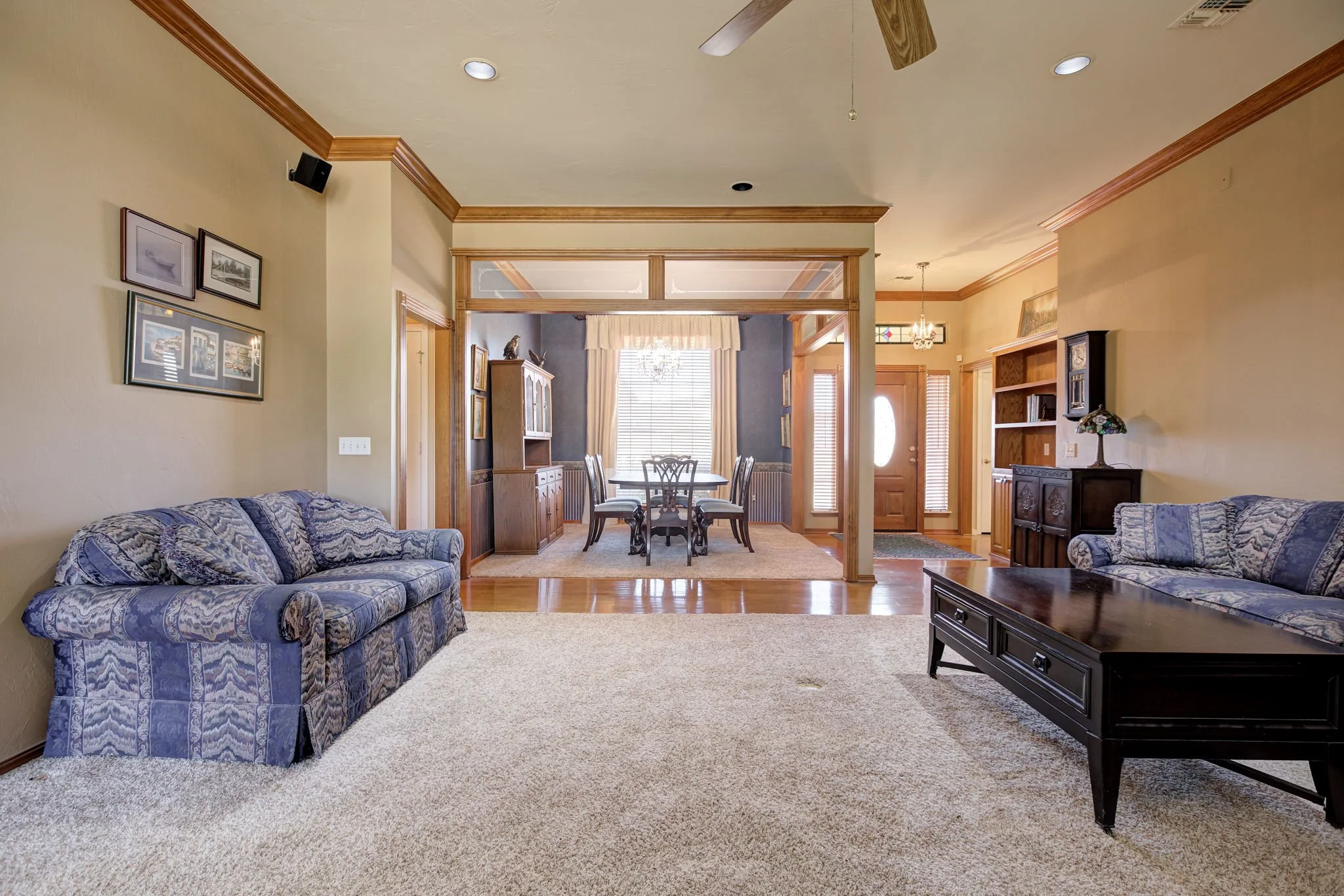 Living room with beige carpet, two blue patterned sofas, black coffee table, wall art, and a view into the dining area with a wooden dining table and chairs, surrounded by windows with blinds and curtains.