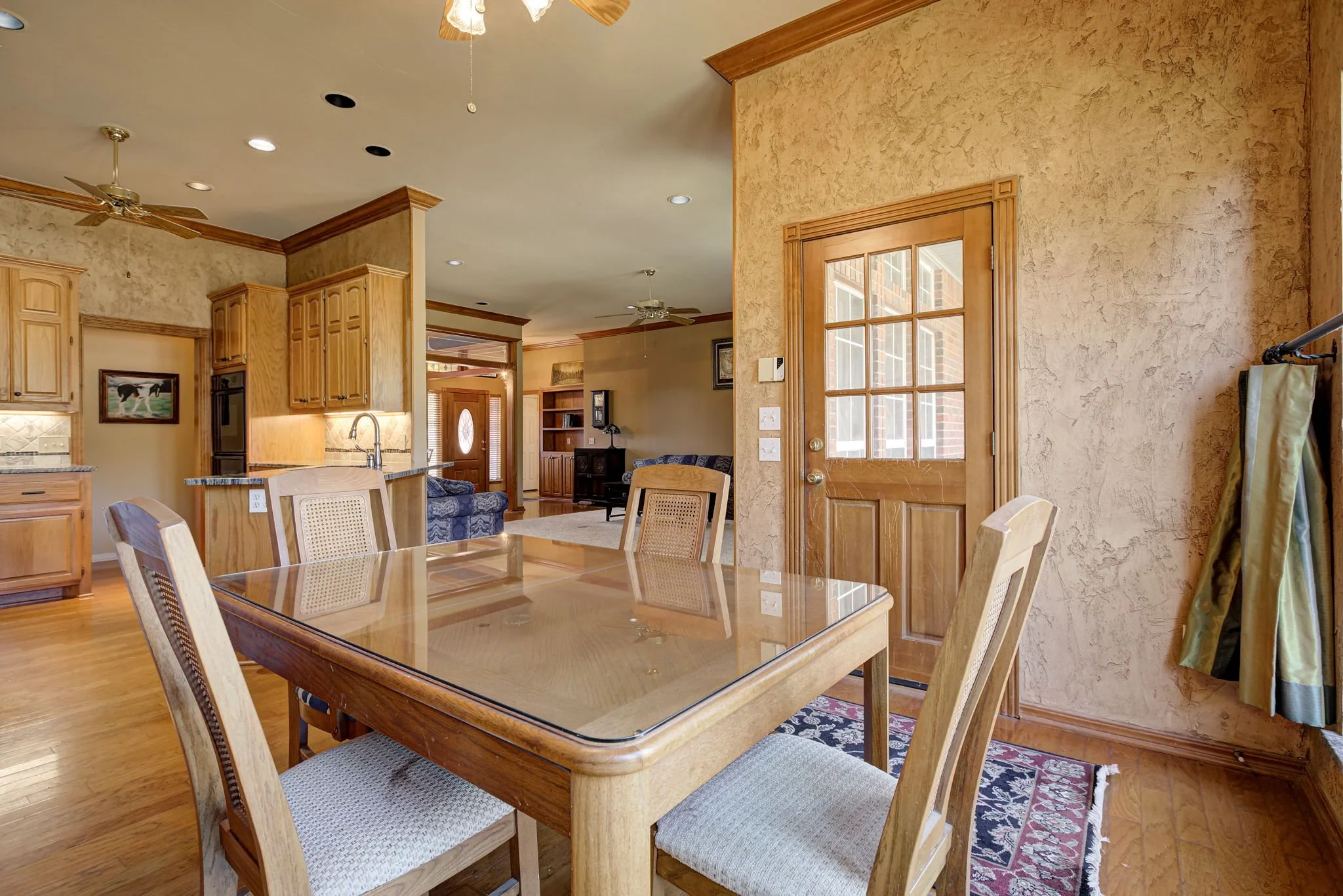 Bright dining and living room with wooden furniture, a glass-top dining table, a wooden door with glass window, and a ceiling fan.