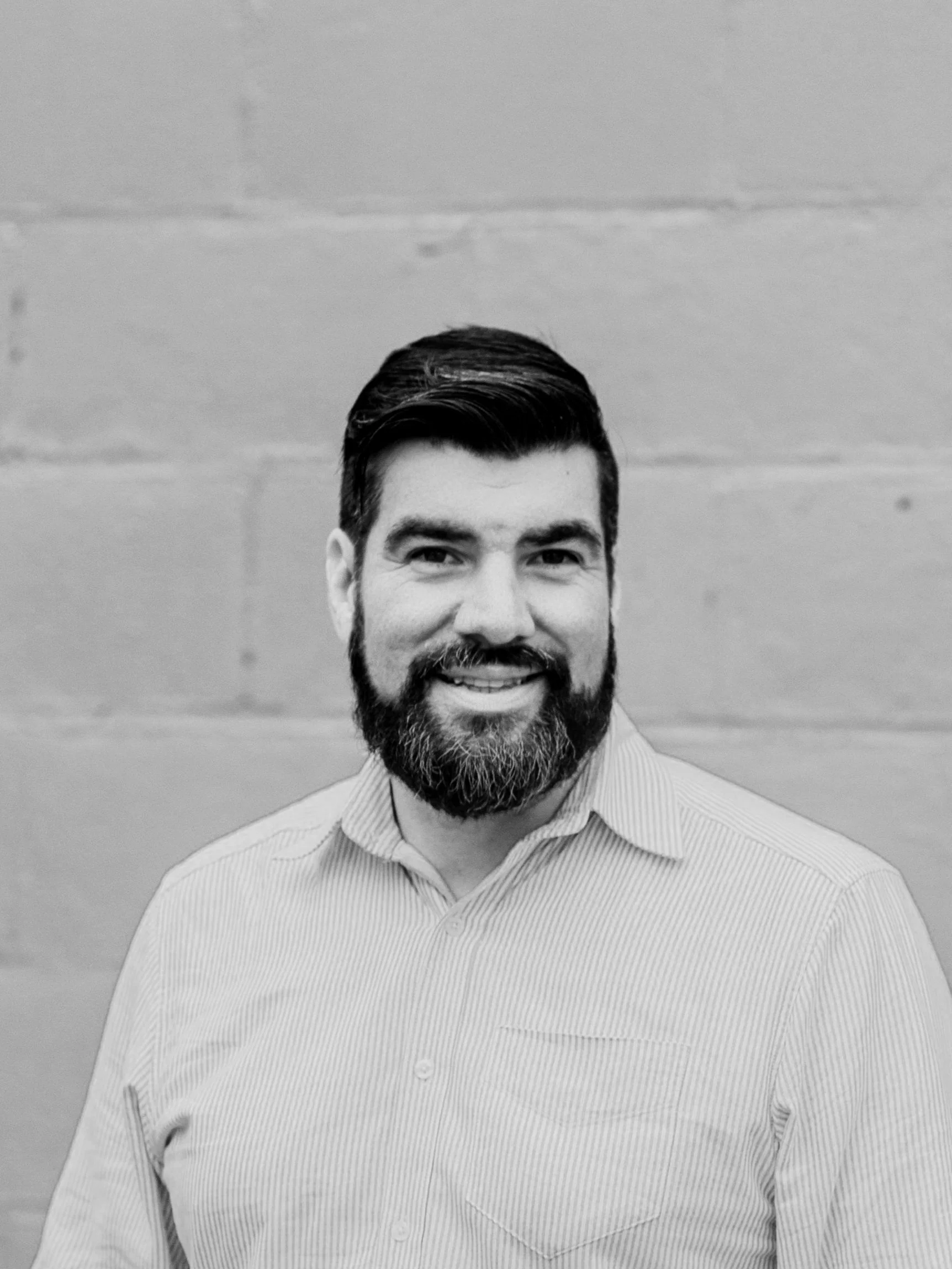 Black and white portrait of a man with dark hair and a beard, smiling, in front of a plain wall.