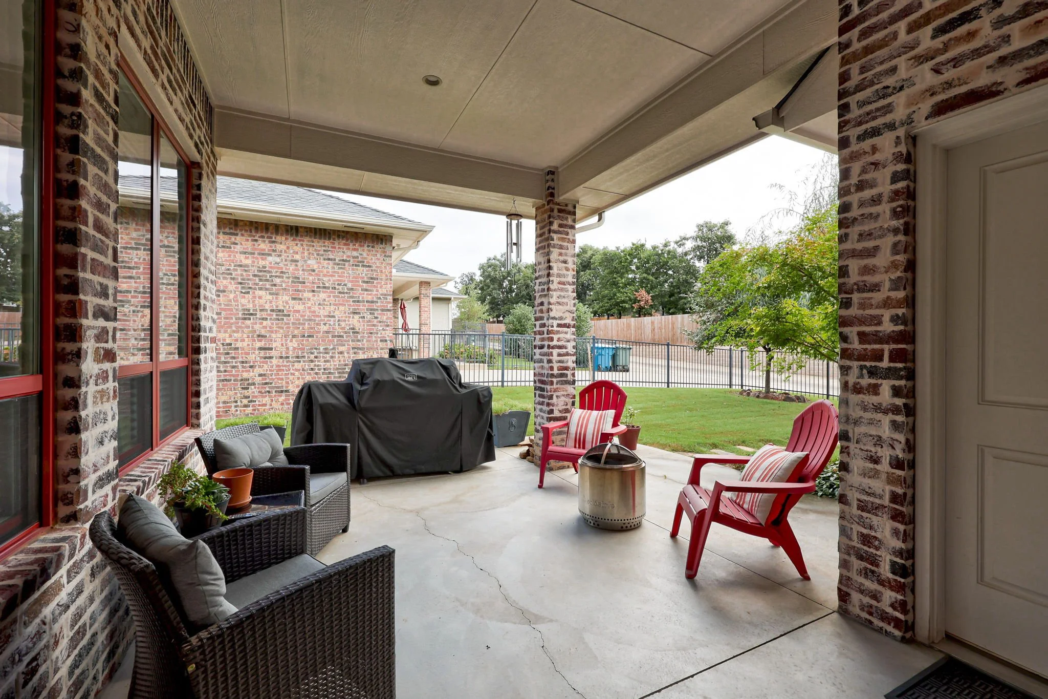 Covered backyard patio with brick columns, wicker chairs, red Adirondack chairs with striped cushions, a grill covered with a black cover, and a metal fire pit, overlooking a fenced grassy yard with trees.
