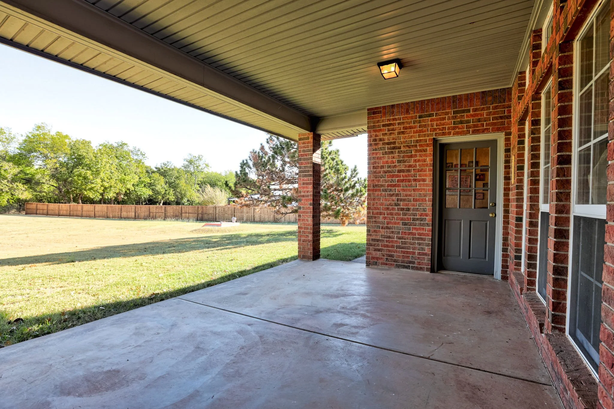 Back porch with concrete floor, brick wall, black door with window, and ceiling light, overlooking backyard with grass, trees, and a wooden fence.