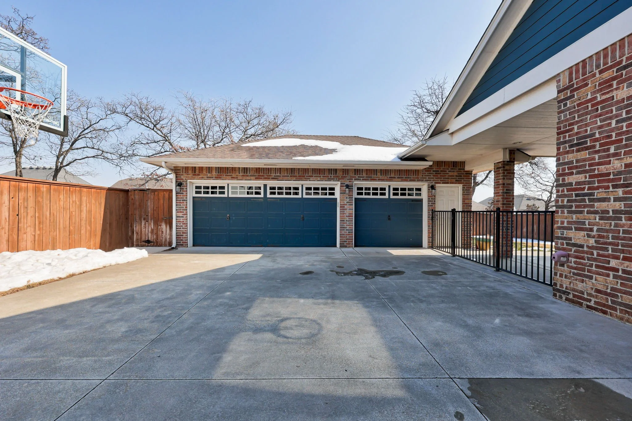 A residential driveway with three blue garage doors and a basketball hoop in the background. The driveway is clean with some wet spots and shadows, a wooden fence on the left, and part of the house with brick walls on the right.