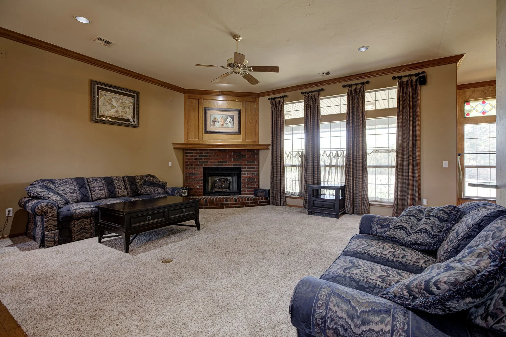 Living room with large windows, patterned sofas, a coffee table, a fireplace with wooden trim, framed art, and a ceiling fan.