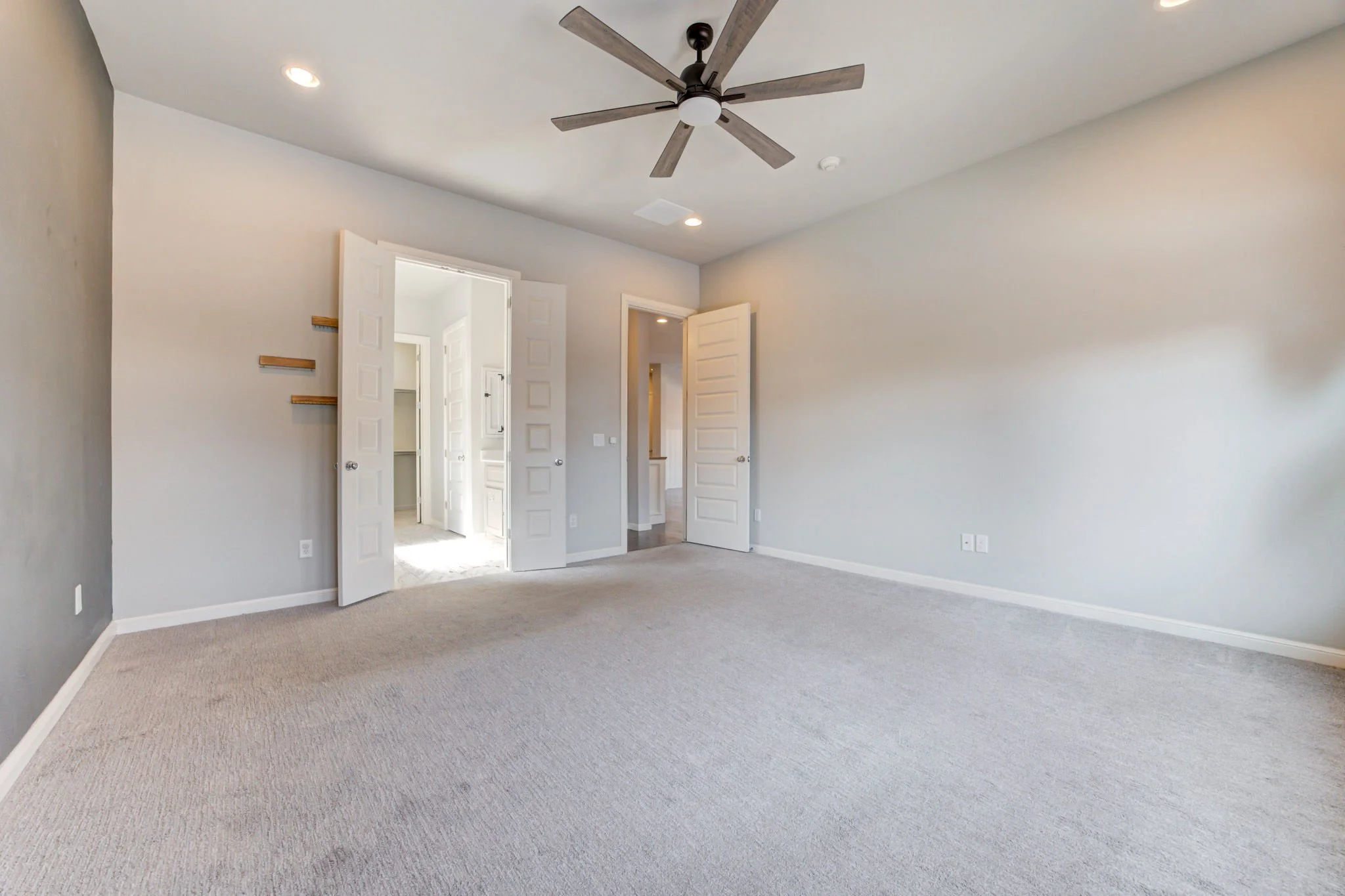 Empty bedroom with light-colored walls, beige carpet, ceiling fan, and open double doors leading to a closet or another room.