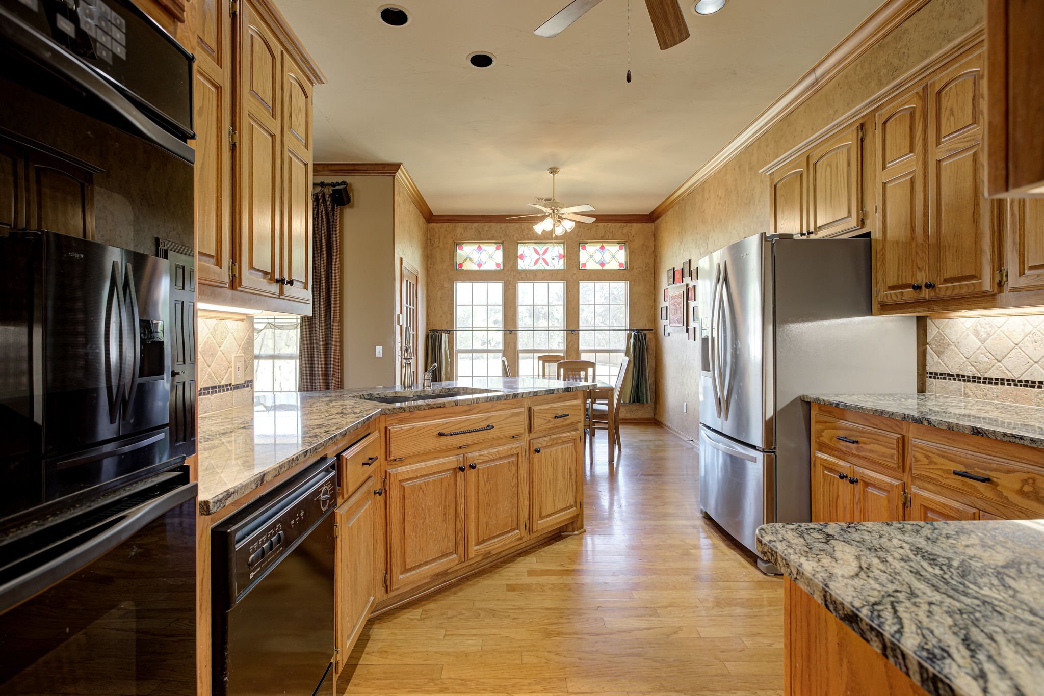 View of a traditional kitchen with wooden cabinets, granite countertops, and stainless steel appliances. There is a breakfast area with large windows and a ceiling fan.