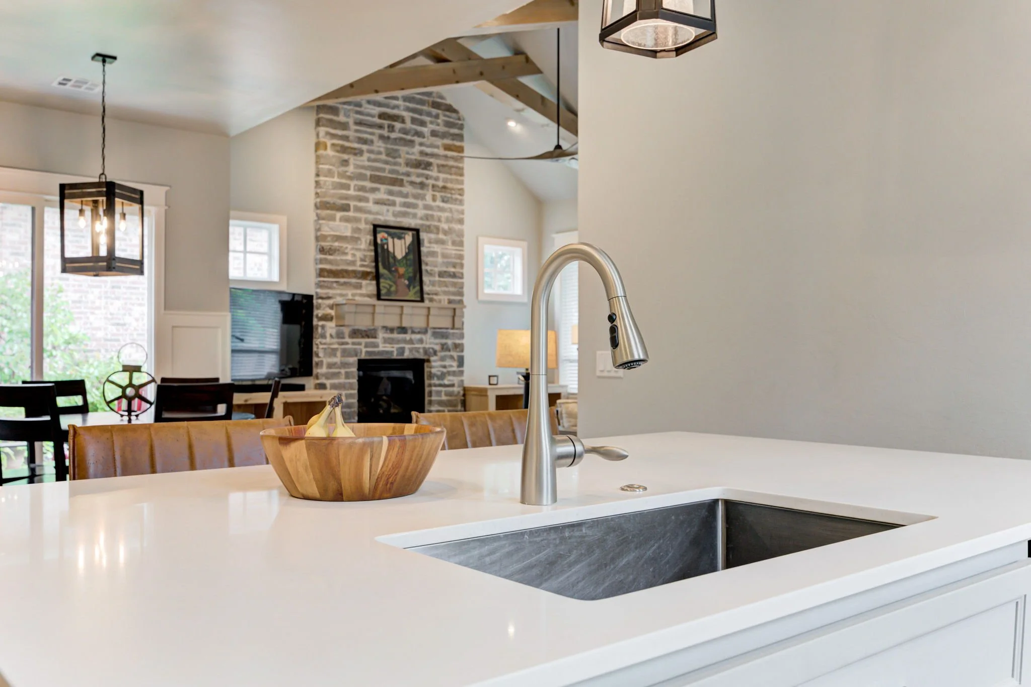 Modern kitchen with white countertops, stainless steel faucet, and a wooden fruit bowl, overlooking a living room with a brick fireplace and dining area.