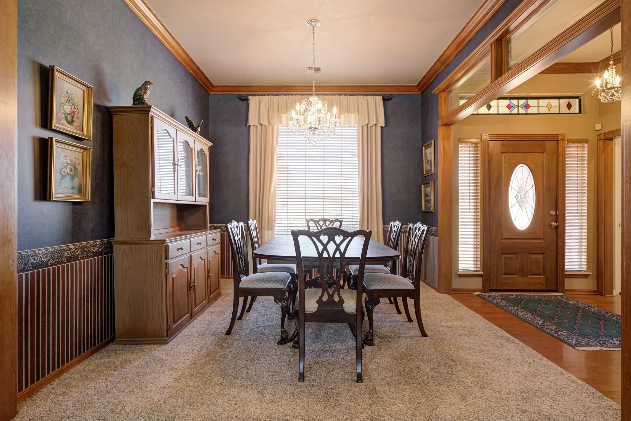 A dining room with a wooden table and six chairs, a wooden cabinet, paintings on the wall, a chandelier, and a large window with blinds and curtains. The front door with an oval glass window is visible on the right.