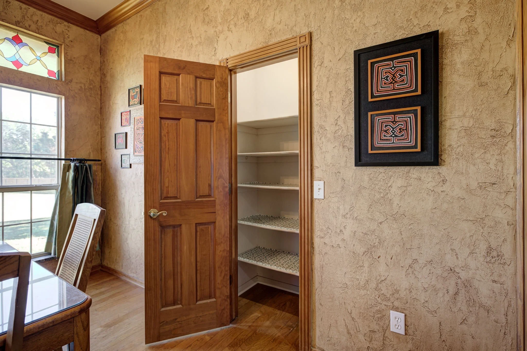 Open wooden door leading to a closet with shelves, beige textured walls, framed artwork, and a large window with stained glass design in a room.