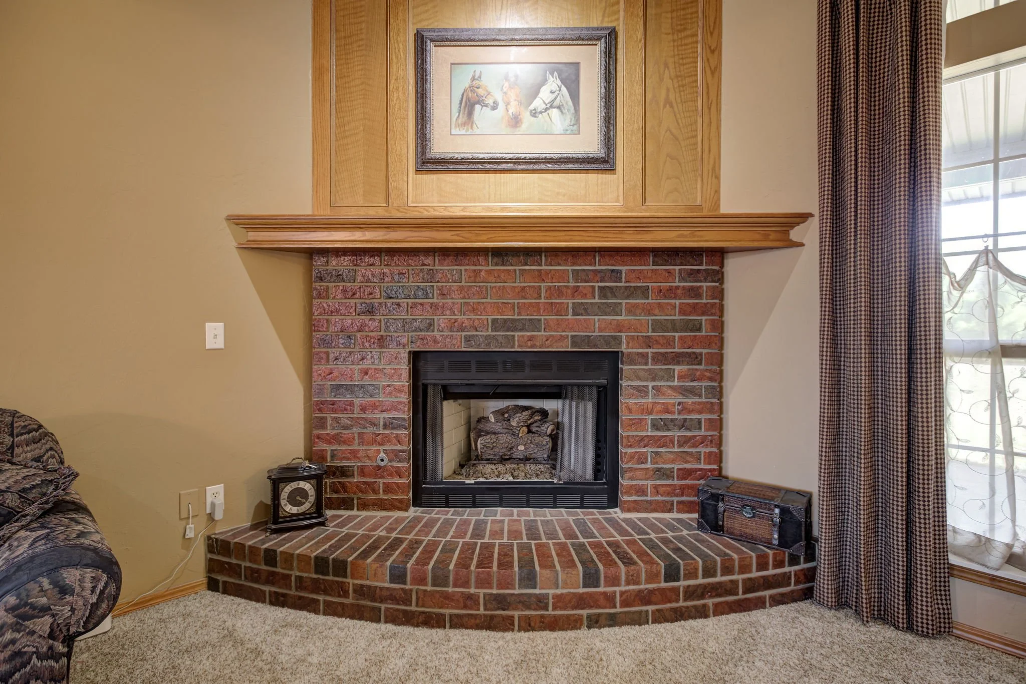 Living room fireplace with a brick hearth, wooden mantel, and framed horse artwork above; to the left a patterned sofa, and to the right a window with checkered curtains.