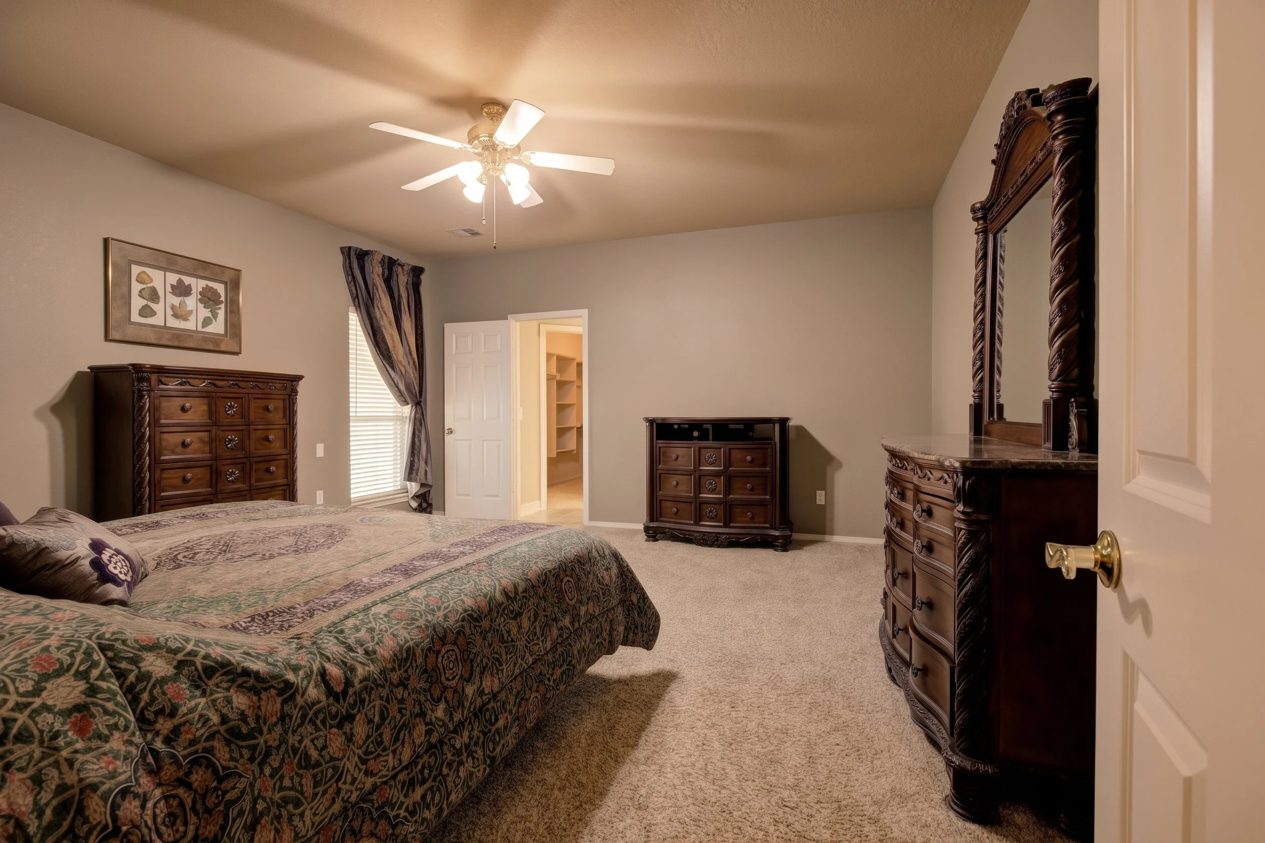 A bedroom with a bed, tall dark wood dresser, matching tall dresser with mirror, and an open door leading to a walk-in closet. The room has a ceiling fan with lights, a window with patterned curtains, and a framed artwork of leaves on the wall.