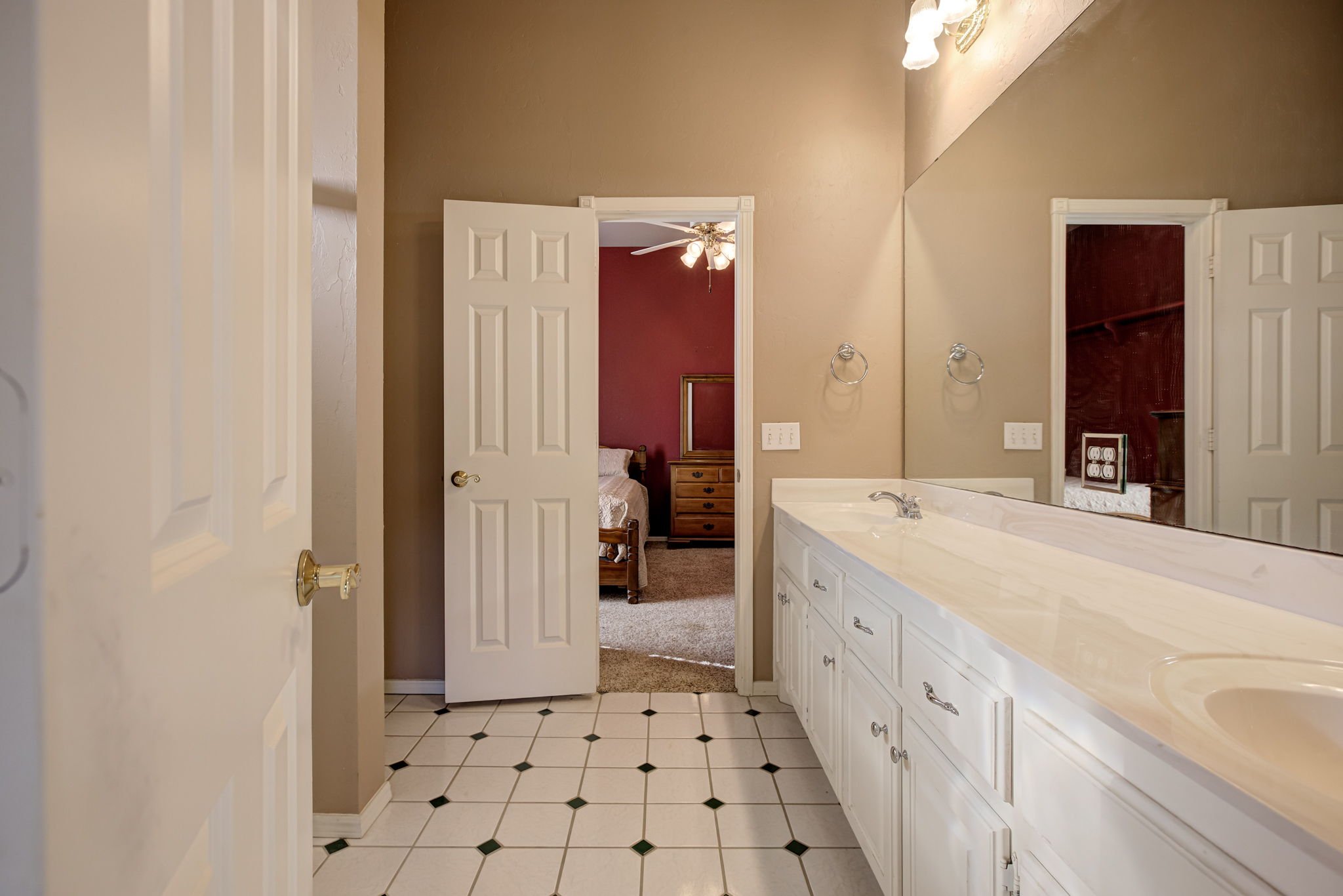 A bathroom with white cabinets, a large mirror, and a vanity sink. There are two open doors leading to a bedroom with a red wall, a ceiling fan, and a bed. The floor has white tiles with black accents.