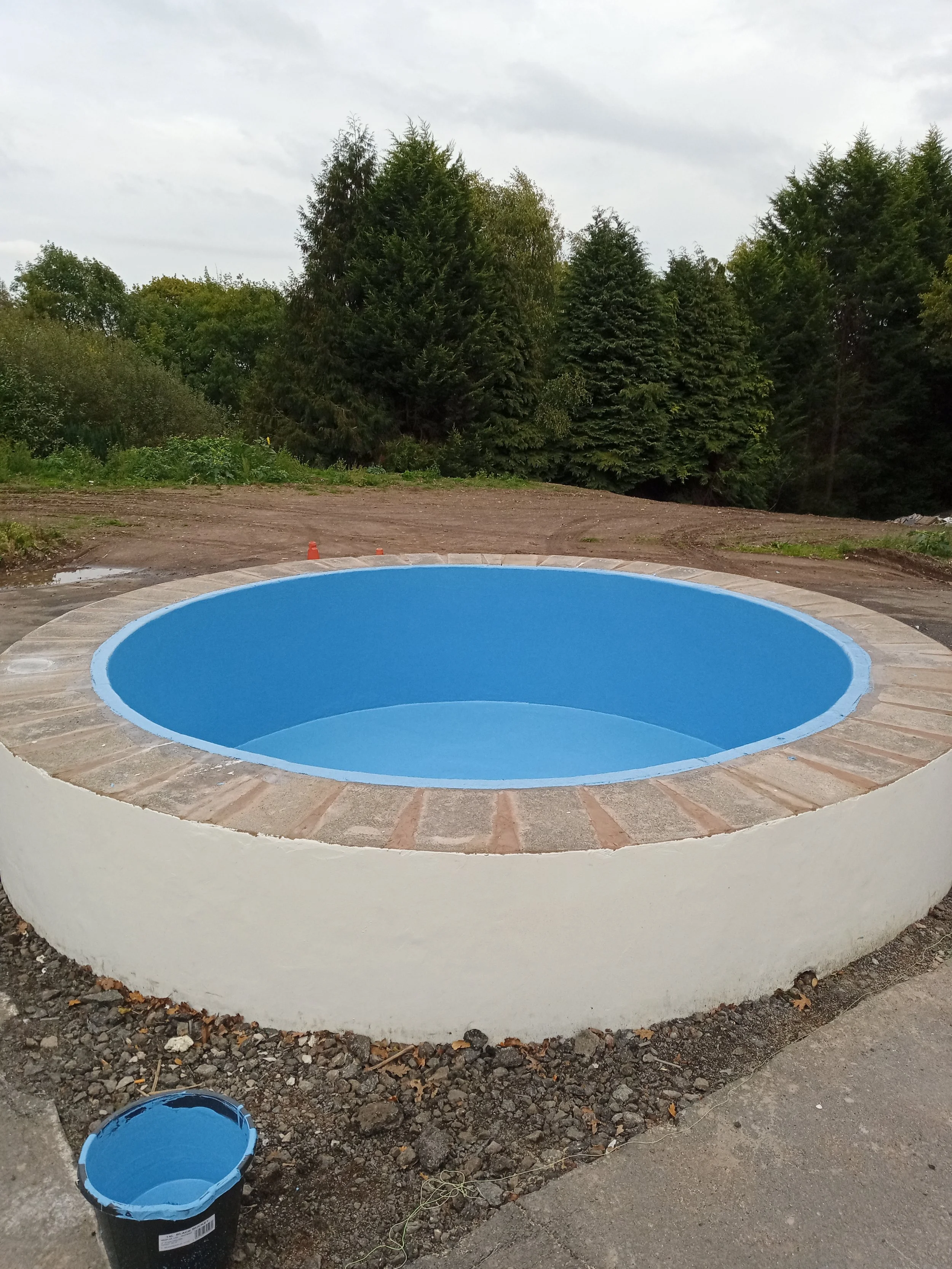 A newly constructed round pool with a white exterior, surrounded by brick coping, and painted blue interior, within a backyard under construction, with trees in the background.