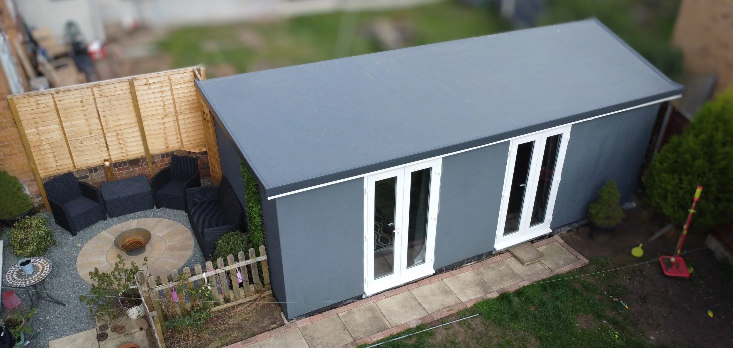 A backyard with a gray shed, a small patio area with black chairs and a fire pit, a wooden fence, and a children's climbing pole on the grass.