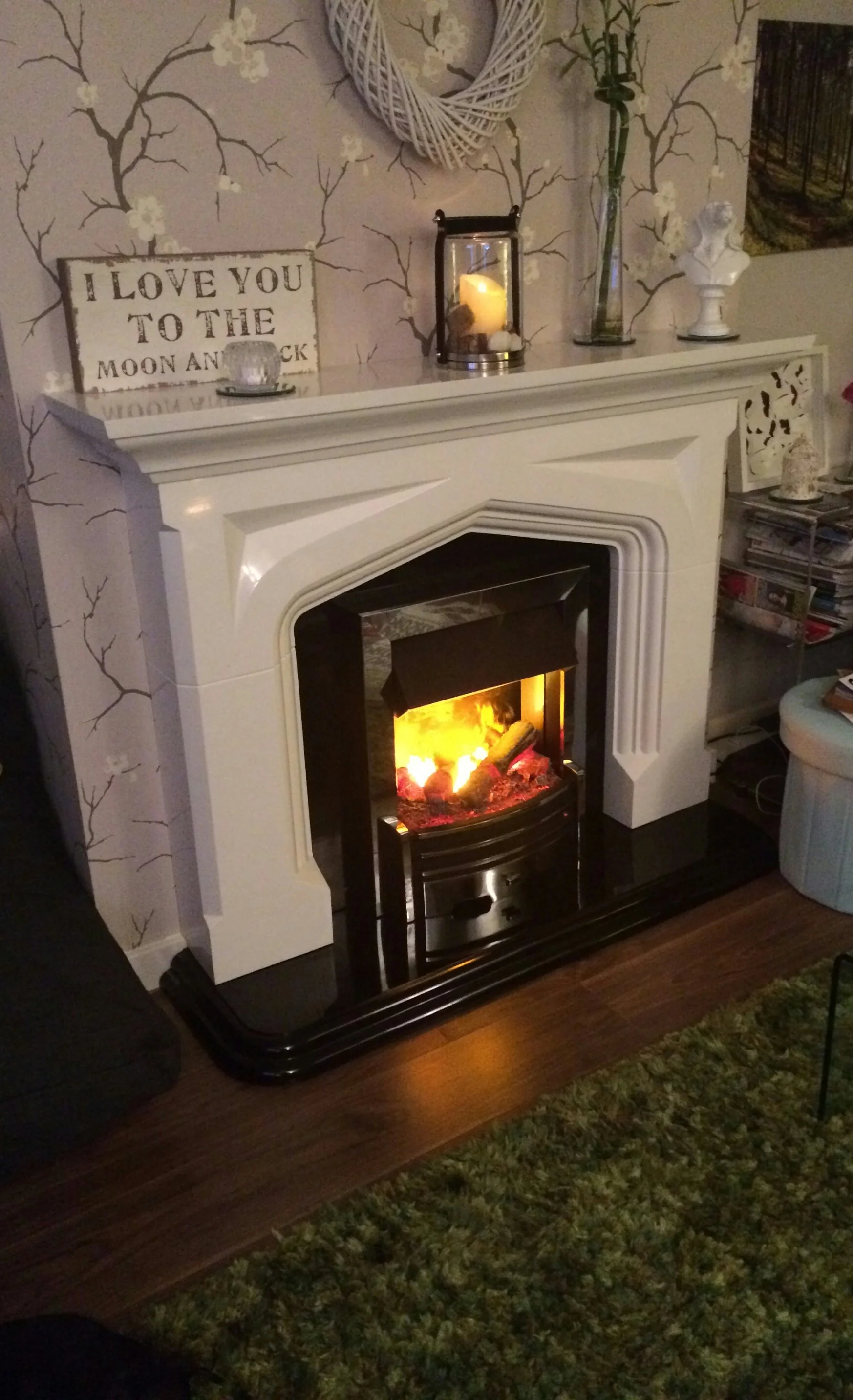 A cozy living room fireplace with a white mantel, decorated with a heart-shaped wreath, a candle lantern, and a vase with flowers. On the mantel, there's a sign that says "I love you to the moon and back." The fireplace has a warm fire burning inside