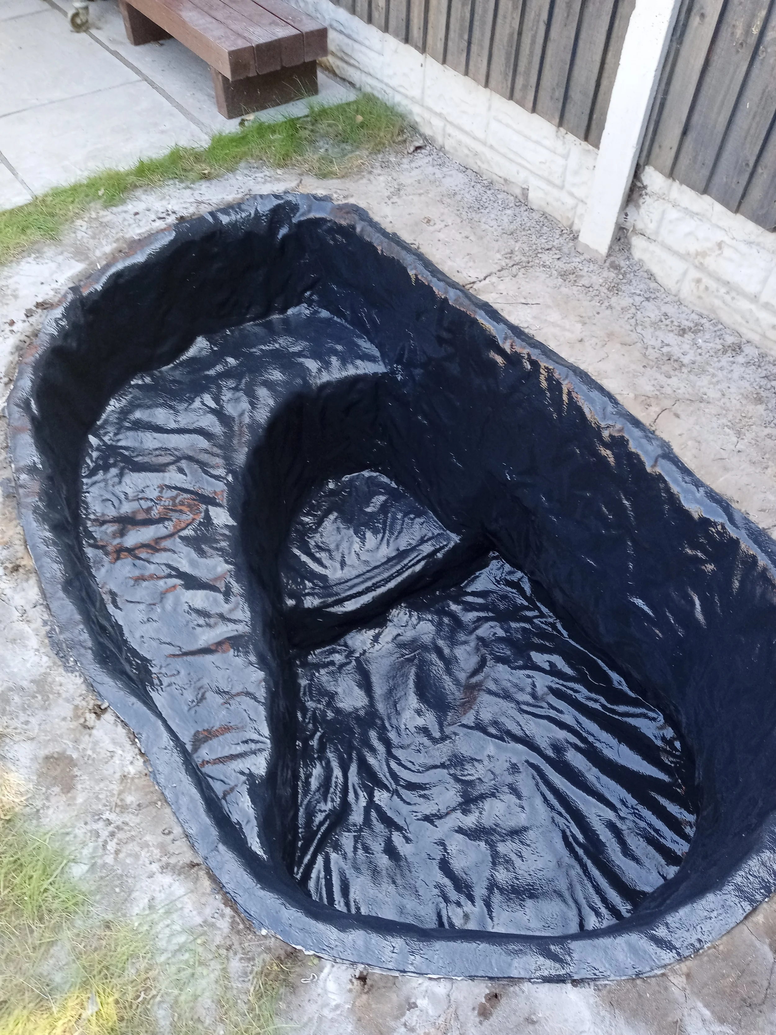 A black plastic pond liner installed in a backyard, forming a pond shape, surrounded by soil, grass, a fence, and a bench.