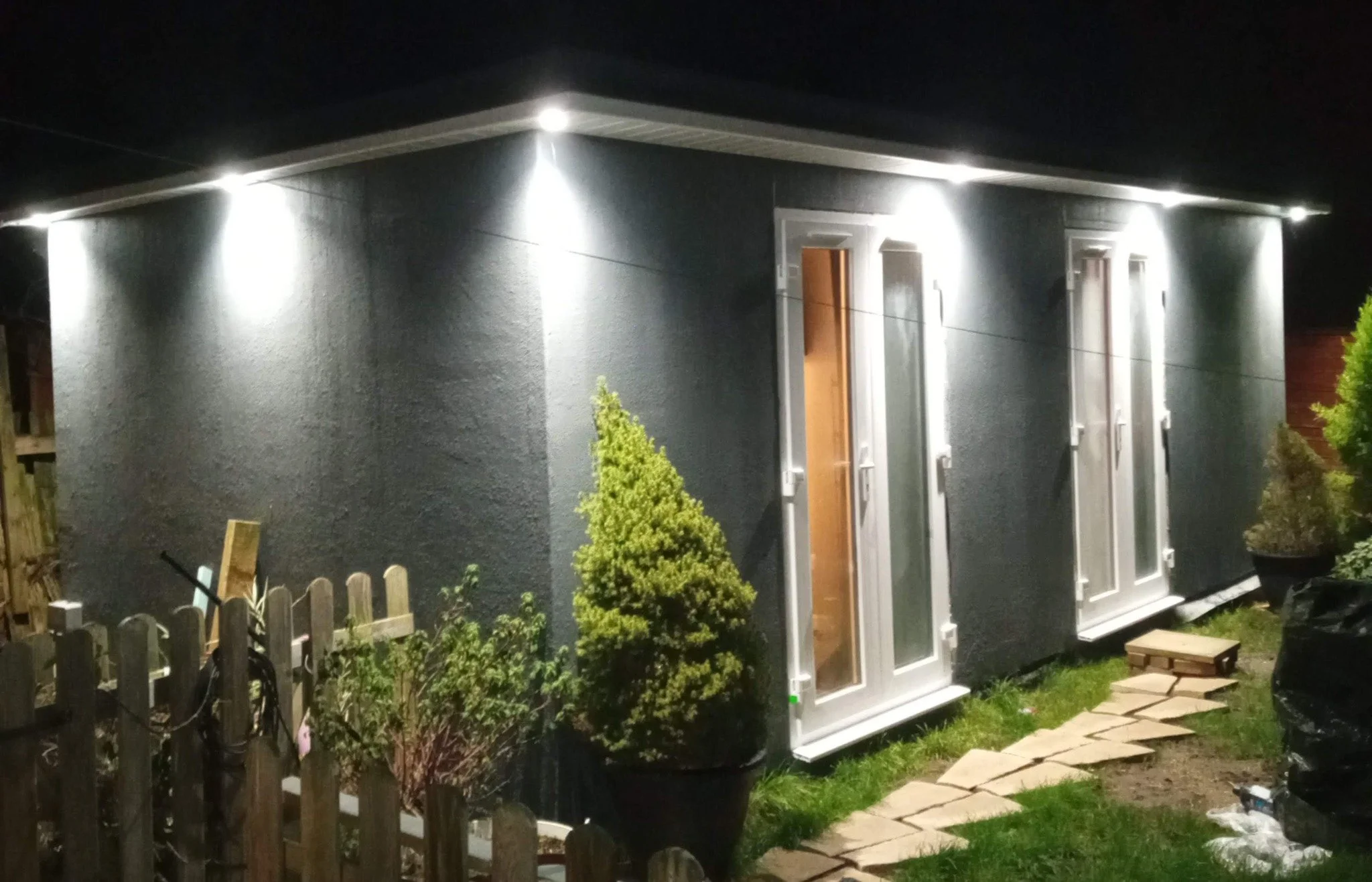 A small backyard house illuminated by exterior lights, with a potted shrub, a wooden fence, and a stone pathway at night.