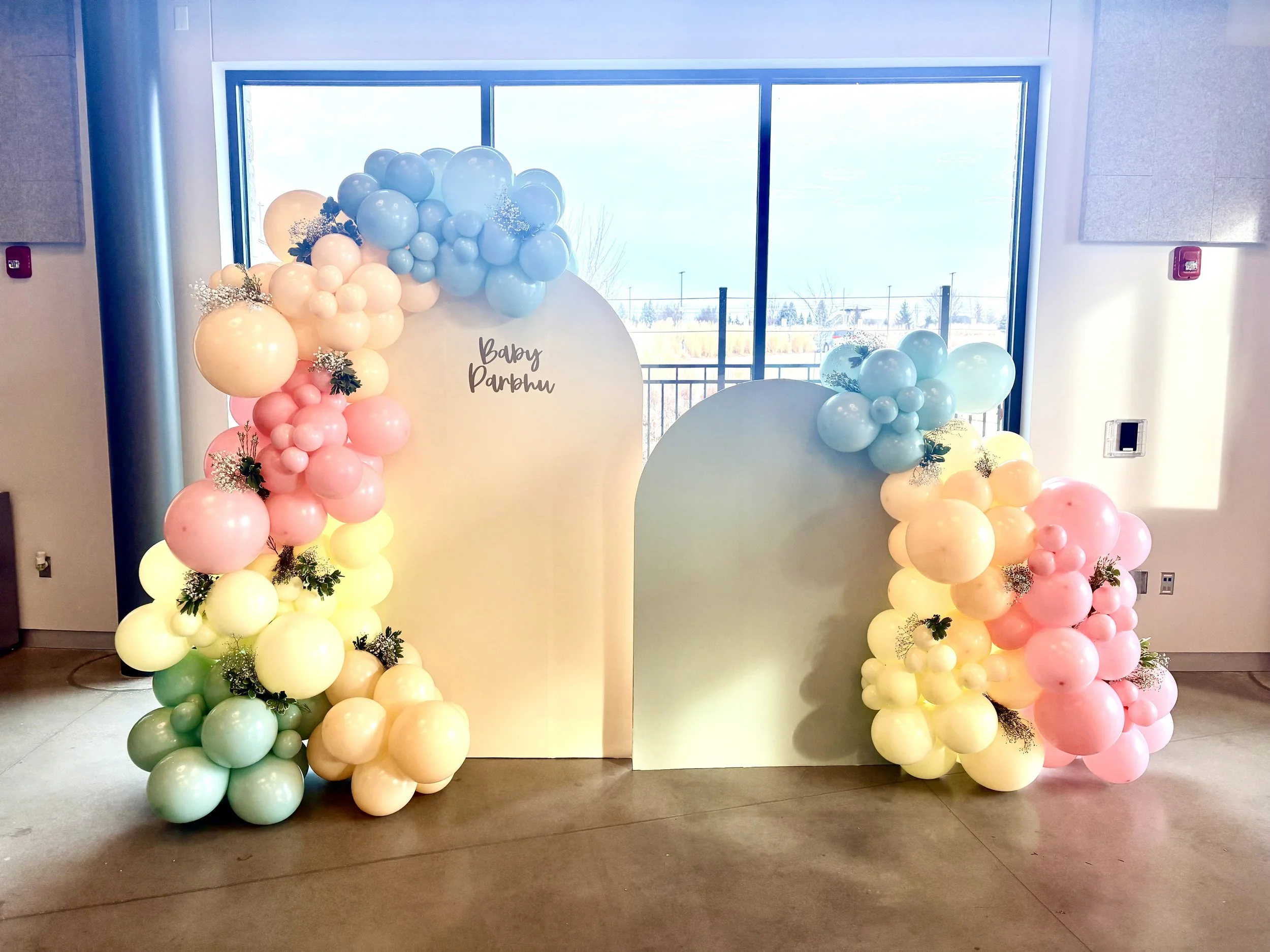 Decorative balloon arch with pastel colors including blue, pink, yellow, and green, featuring baby-themed signage that reads 'Baby Darbhu' in front of a large window.