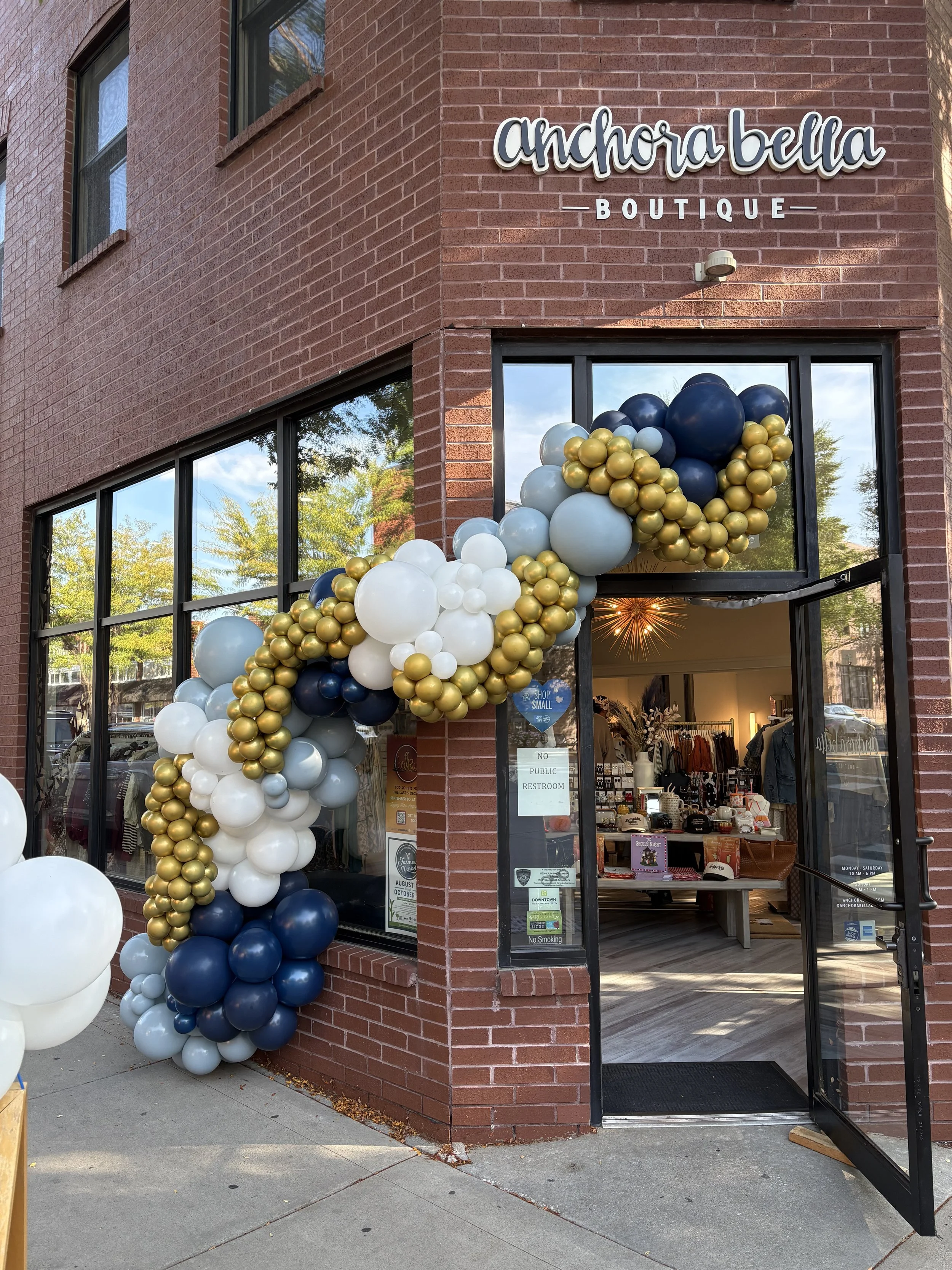 Decorative balloon arch made of white, navy blue, gold, and light gray balloons outside a boutique shop called Anchora Bella.