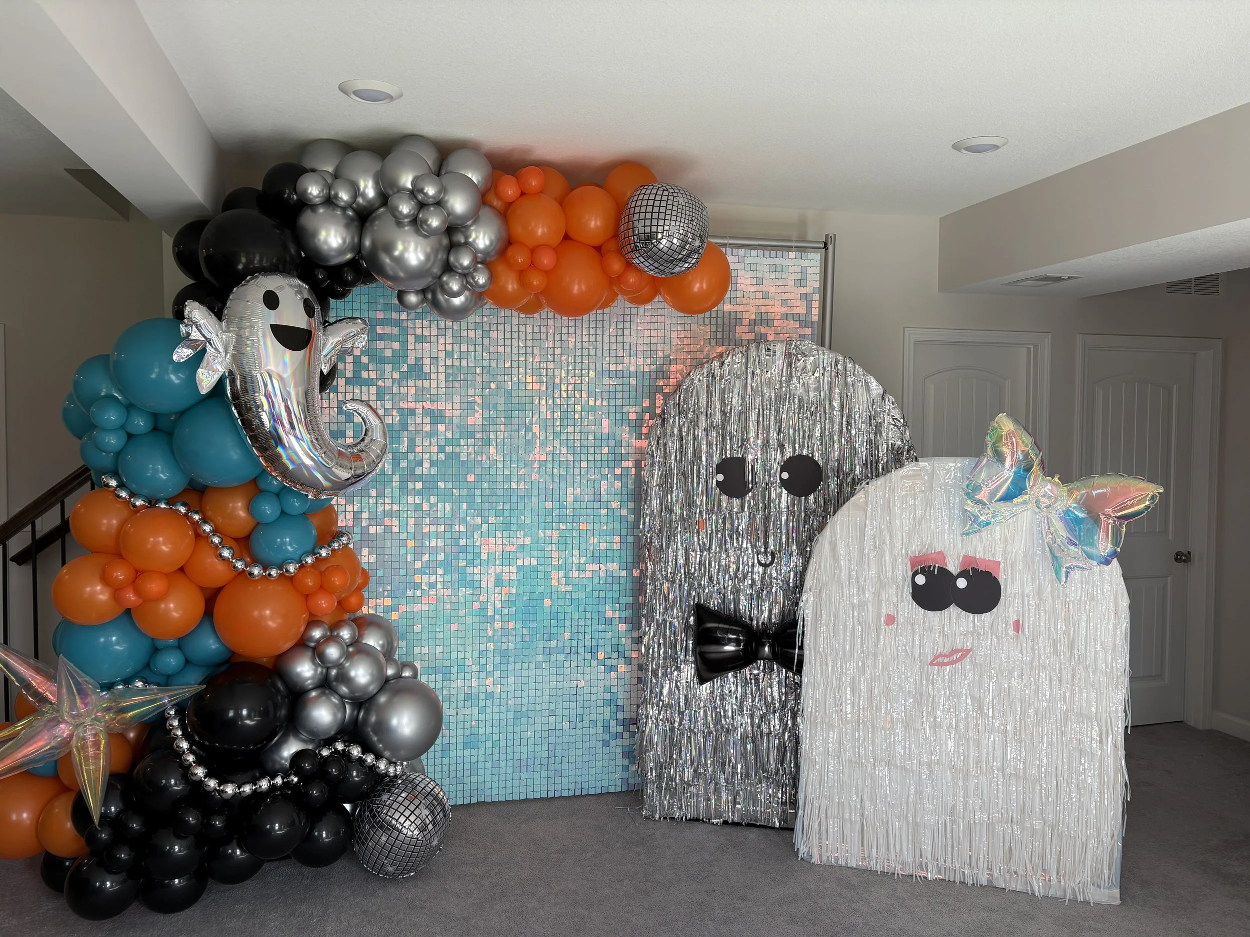 Decorative Halloween display with balloon ghosts, birthday-themed background with balloons and a mosaic wall, foil fringe curtains, and colorful balloon figures with faces, bow, and makeup.