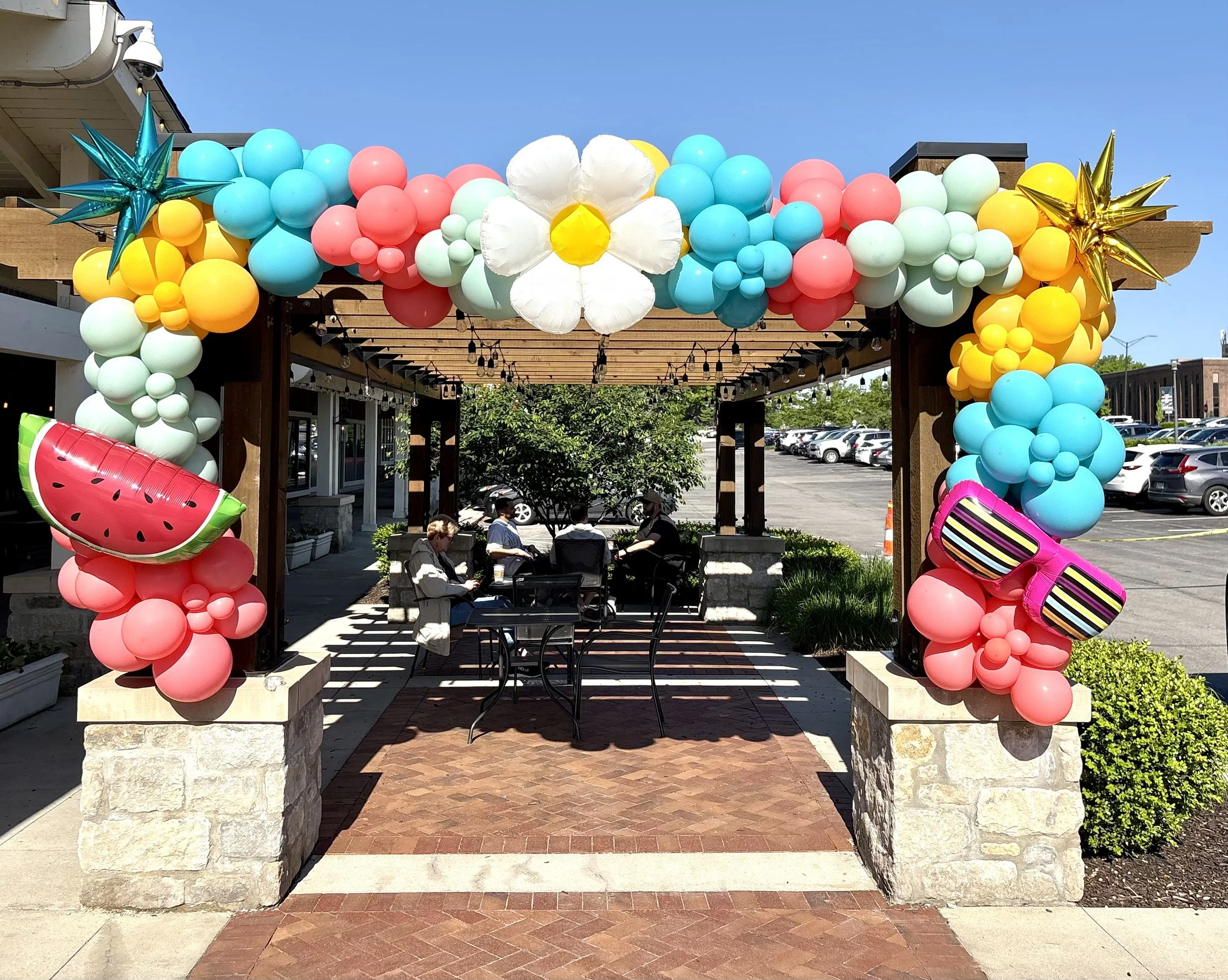 Outdoor dining area decorated with colorful balloon arch featuring strawberries, sunglasses, and a large flower at the top center. People are sitting at tables in the background.