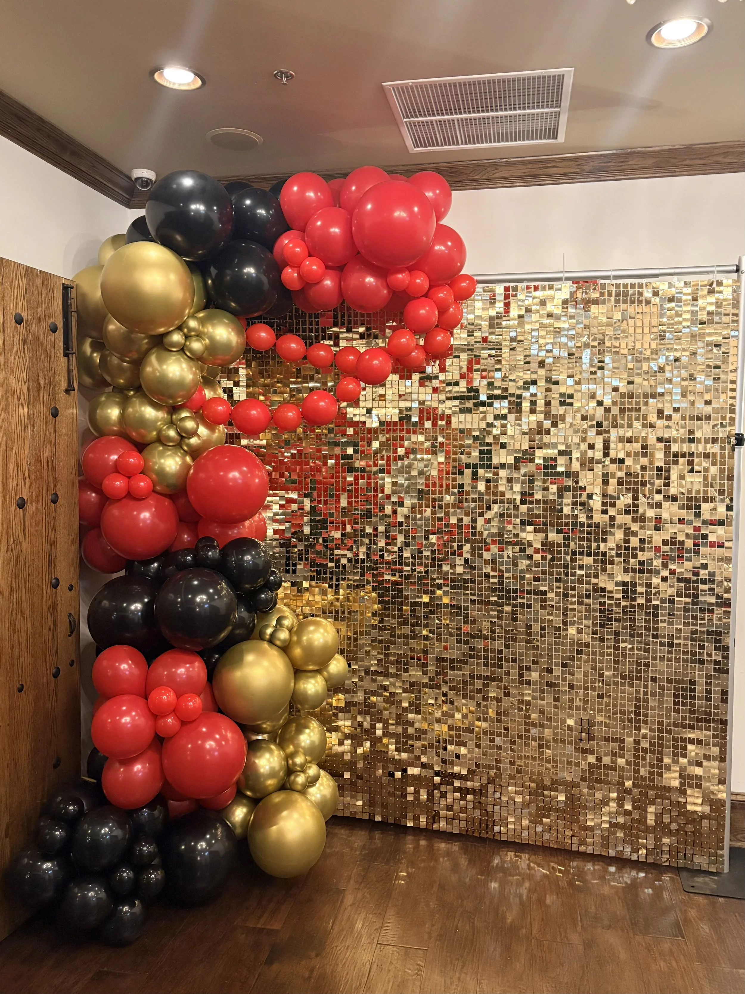 Decorative photo backdrop with red, gold, and black balloons arranged on the left side of a gold sequin curtain backdrop in an indoor setting.