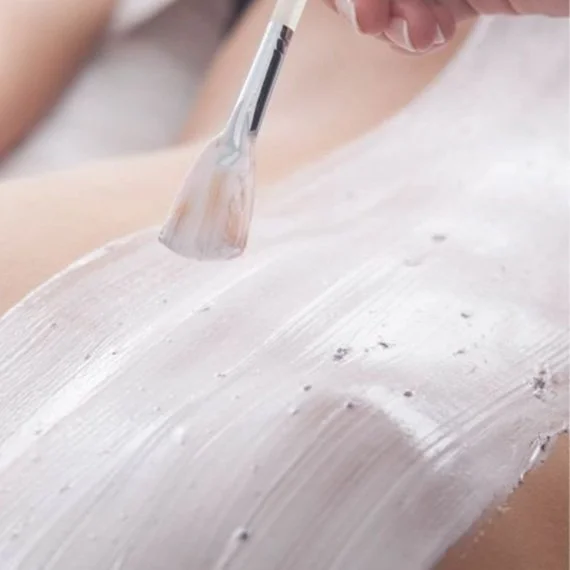 Close-up of a person's body with a brush applying a white mud or clay mask during a spa treatment.