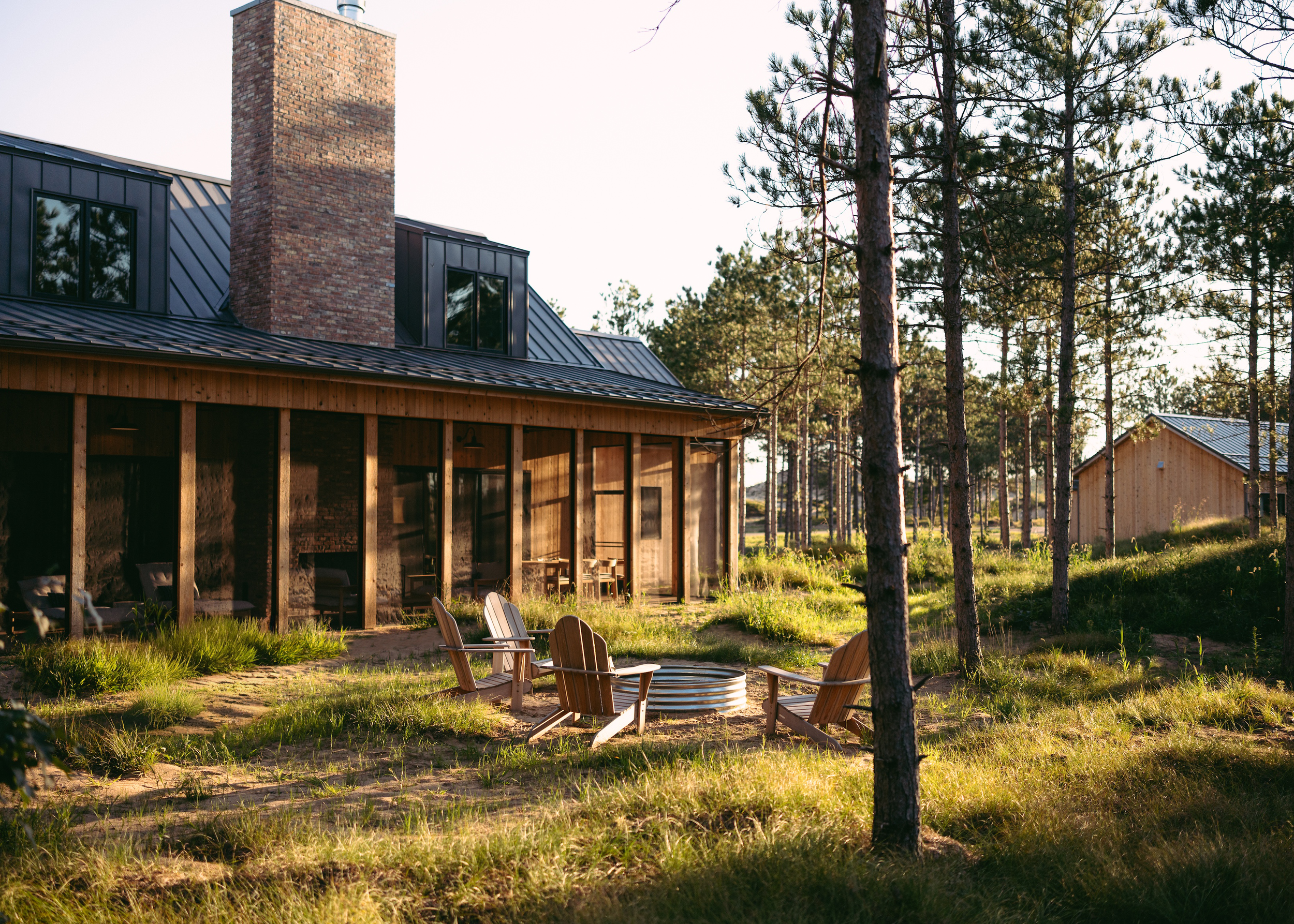 A house with a brick chimney, black metal roof, and wooden exterior walls is situated among tall pine trees. There are Adirondack chairs around a fire pit in the yard, and the sun is setting, casting a warm light.