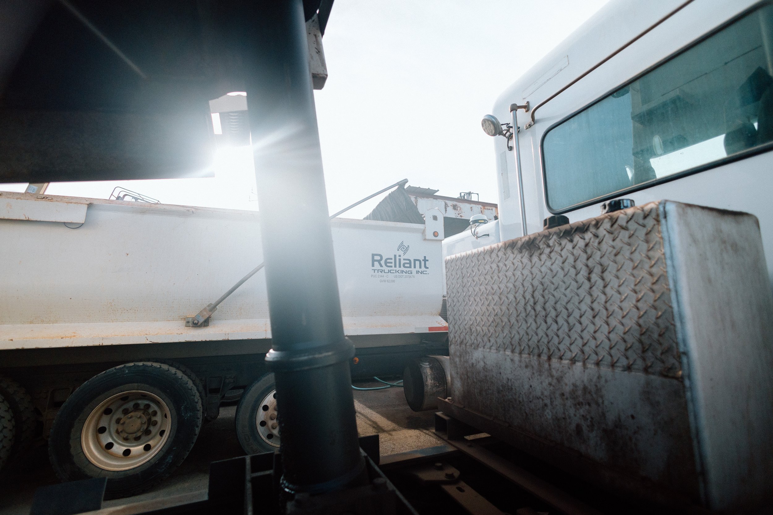Close-up of industrial trucks with a bright sun glare in the background, showing tires, a metal toolbox, and various equipment.
