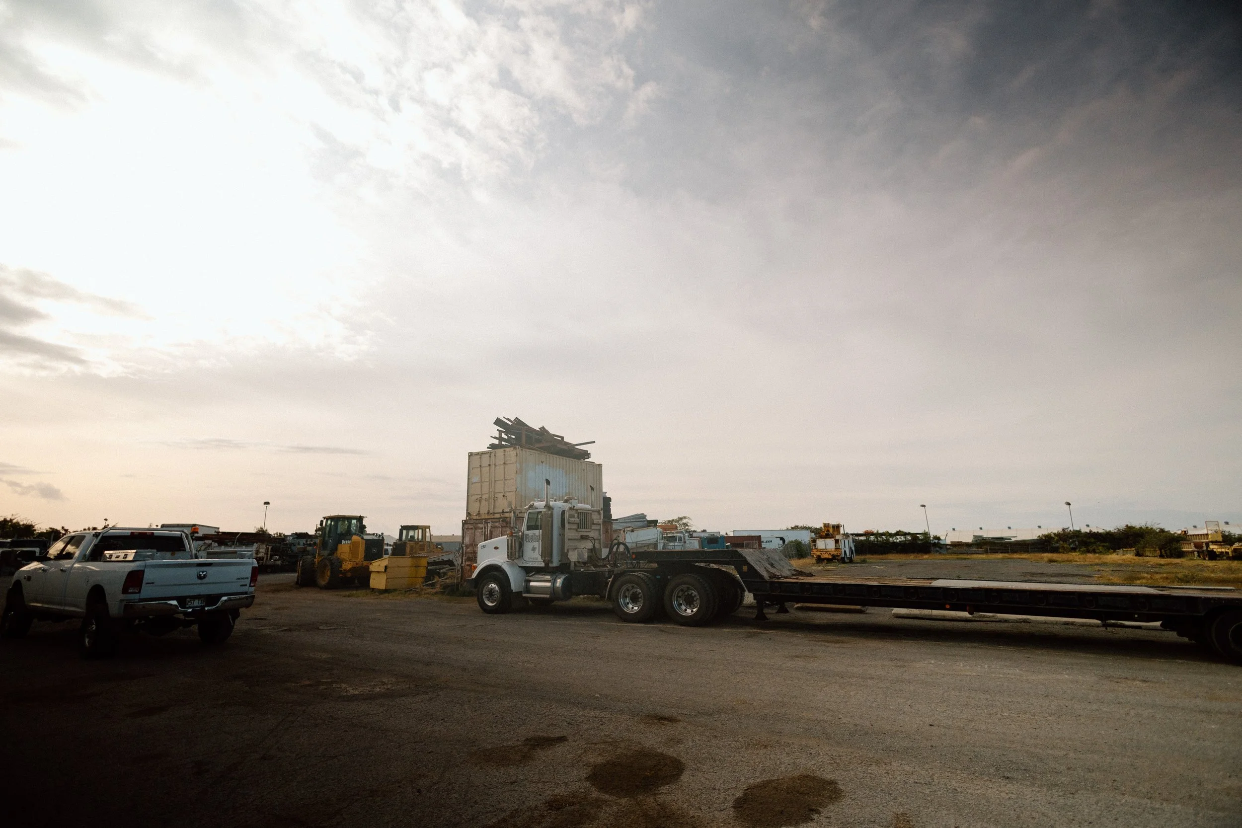 A parking lot with various trucks and heavy machinery under a cloudy sky, including a flatbed truck and a white pickup truck.