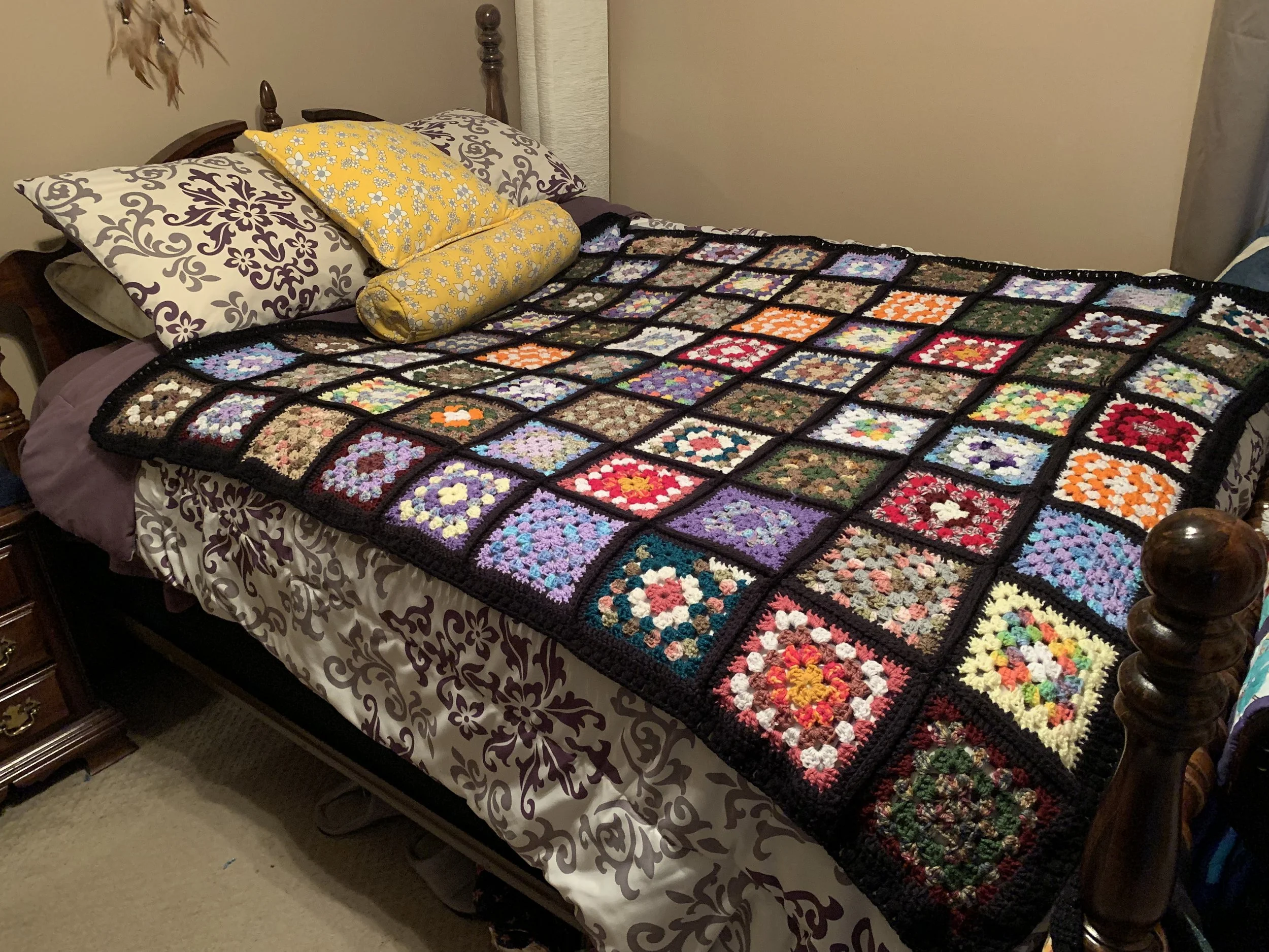 A bed with a handmade patchwork quilt featuring multiple colorful square crochet patterns, two pillows with purple floral covers, and a yellow bolster pillow, in a bedroom with beige walls and a brown dresser.