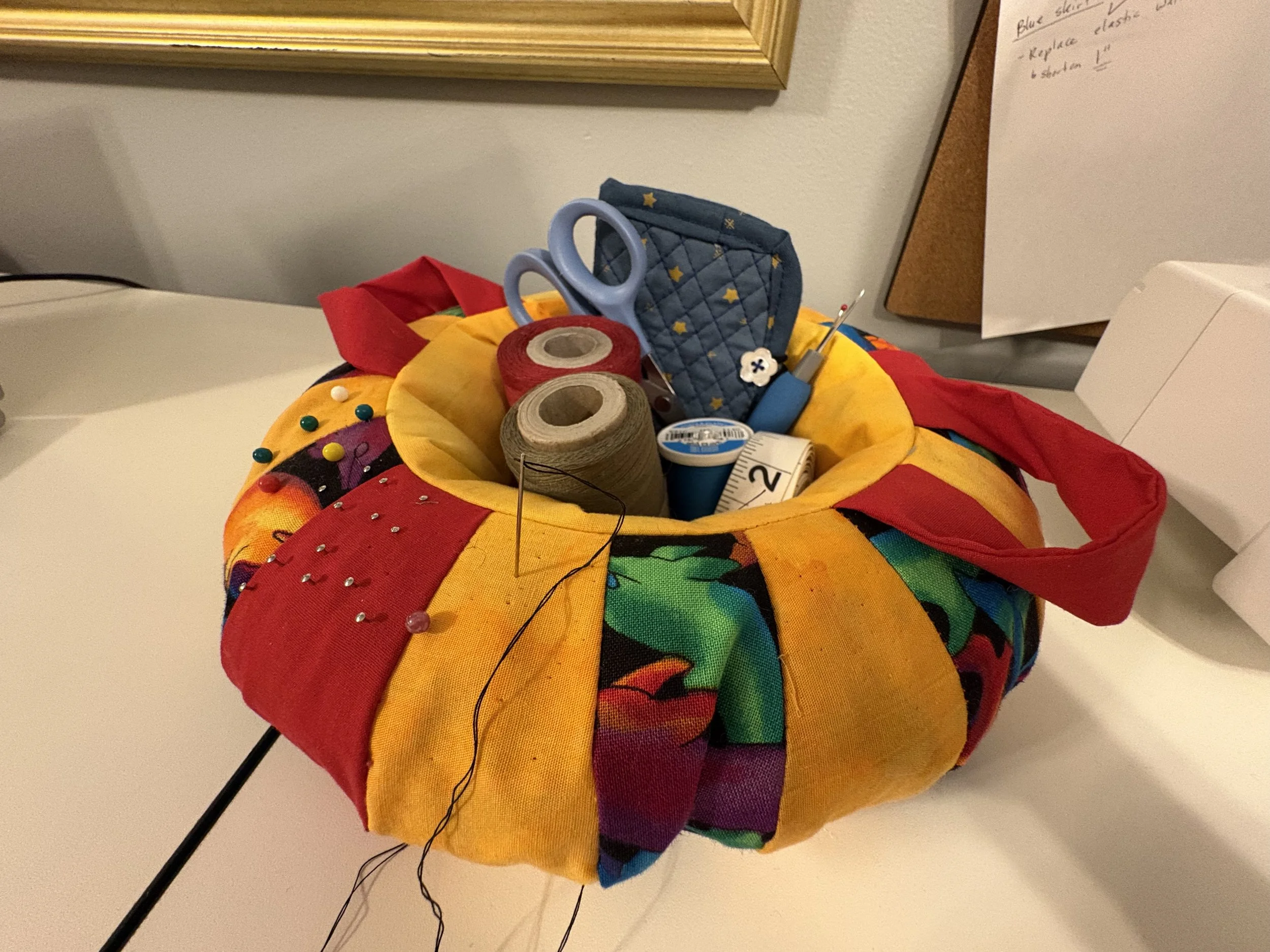 A colorful pin cushion filled with sewing supplies including thread, scissors, measuring tape, pins, and a seam ripper.