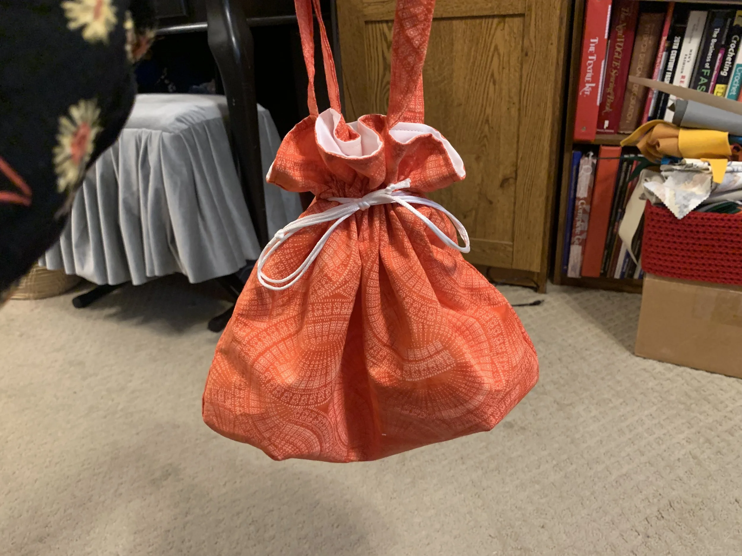 A red gift bag hanging on a hook, with a white ribbon tied around it, in a room with a bookshelf, boxes, and a table.