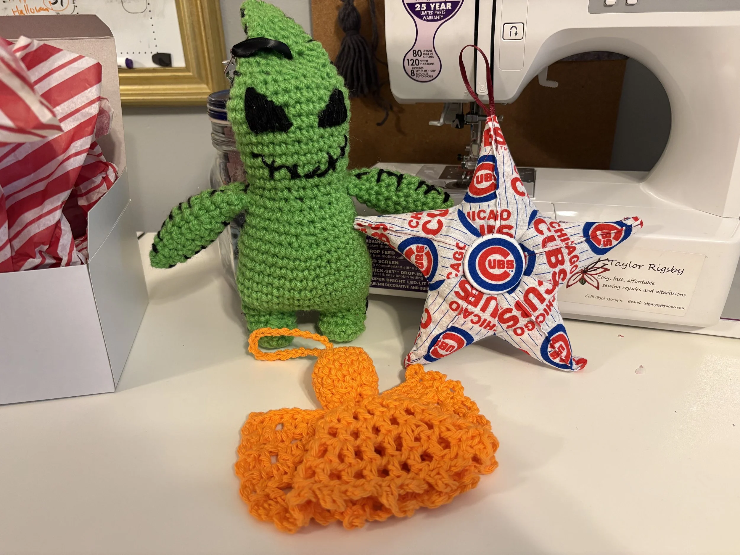 A green crocheted plush toy with black facial features, a hat, and orange feet, standing next to a colorful crocheted star with a Chicago Cubs logo, and an orange crocheted pumpkin on a table with sewing and craft supplies in the background.