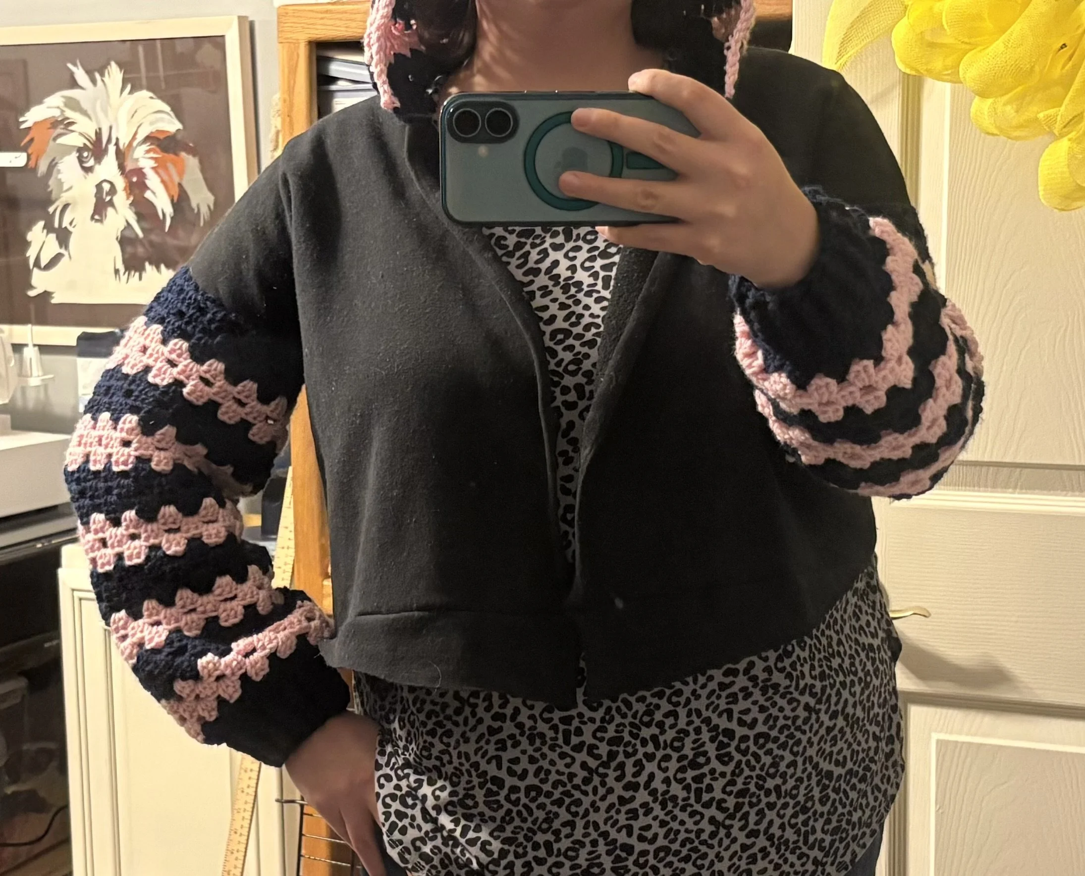 Person taking mirror selfie wearing a black jacket with pink and navy crocheted striped sleeves and a leopard print top and pants in a room with dog art on the wall.