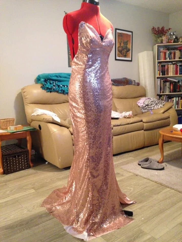 A pink, sequined, strapless evening gown displayed on a red dress form in a living room.