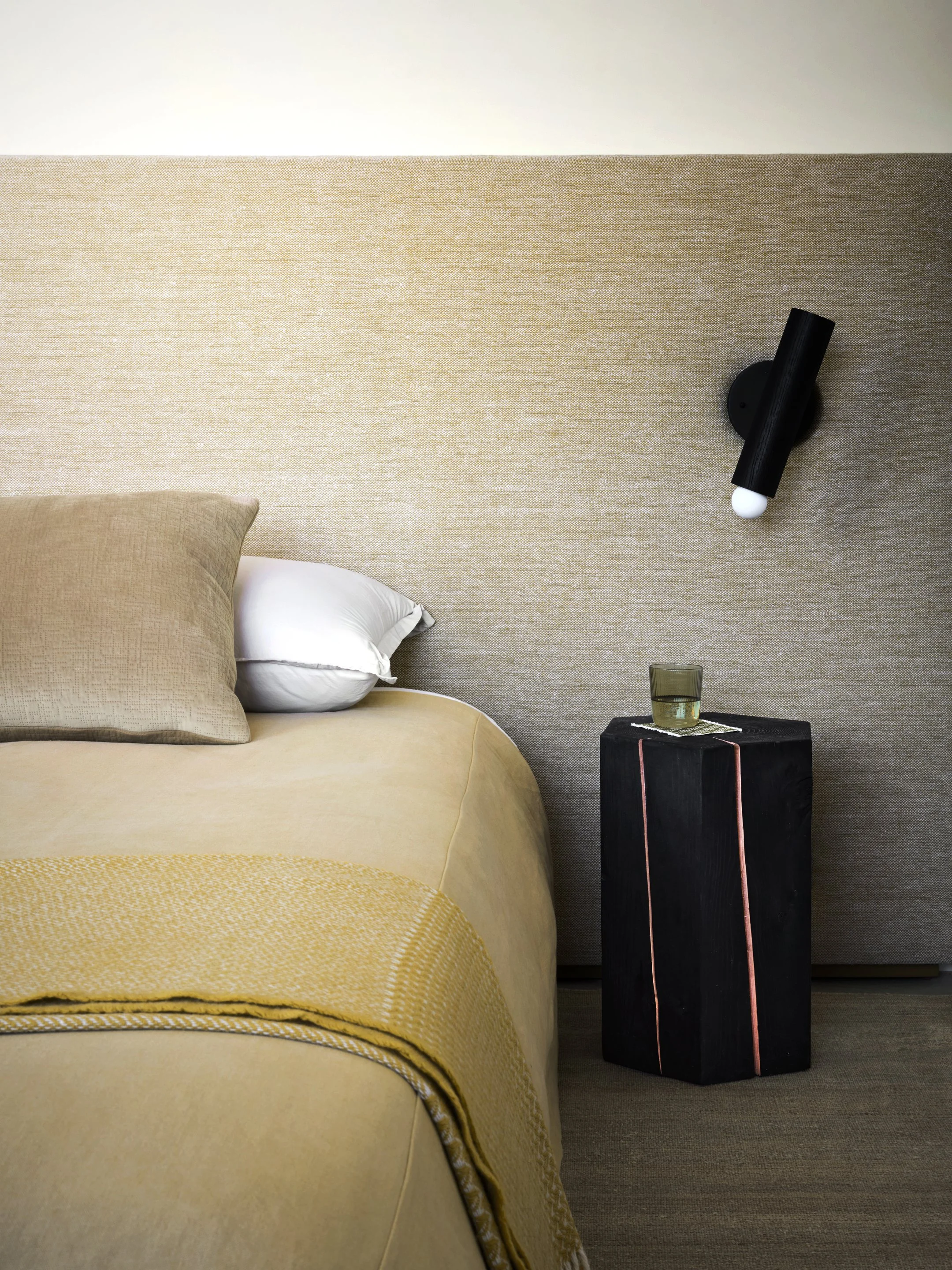 Minimalist bedroom detail with upholstered headboard, wall mounted lighting, and neutral textures