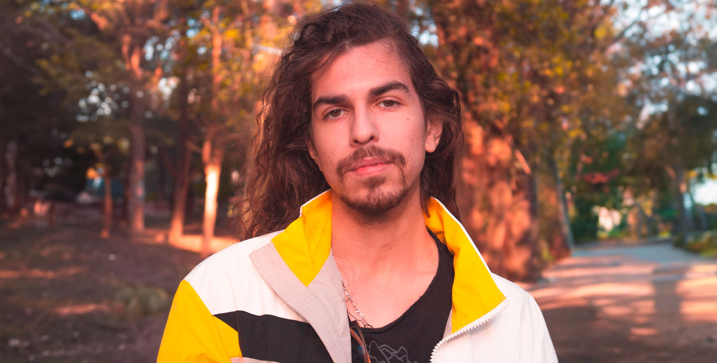 Homem com cabelo longo e barba, usando jaqueta branca com detalhes amarelos, em parque ao pôr do sol