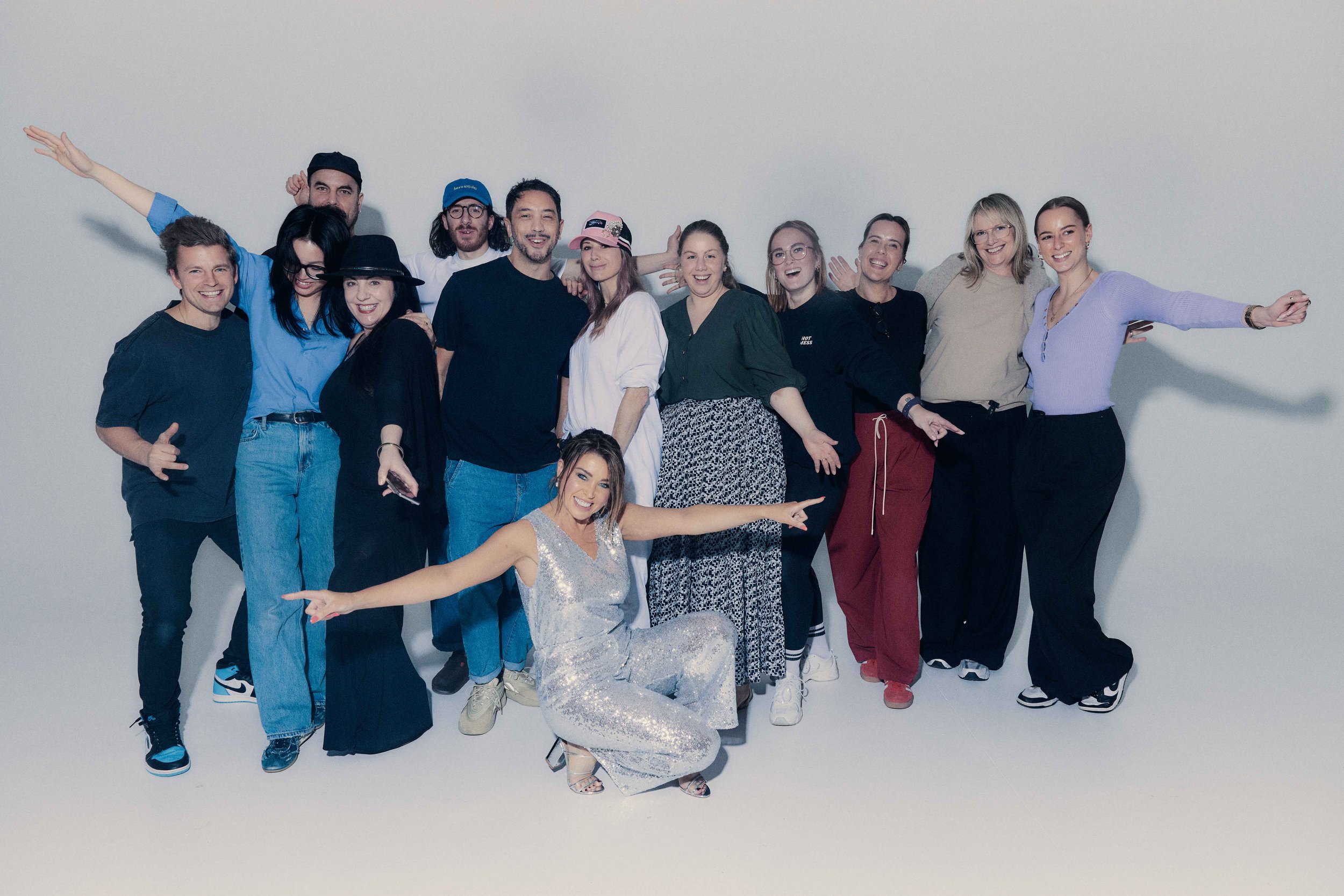 Group of twelve people posing and smiling for a photo against a plain white background, with one woman kneeling in front pointing to the side.