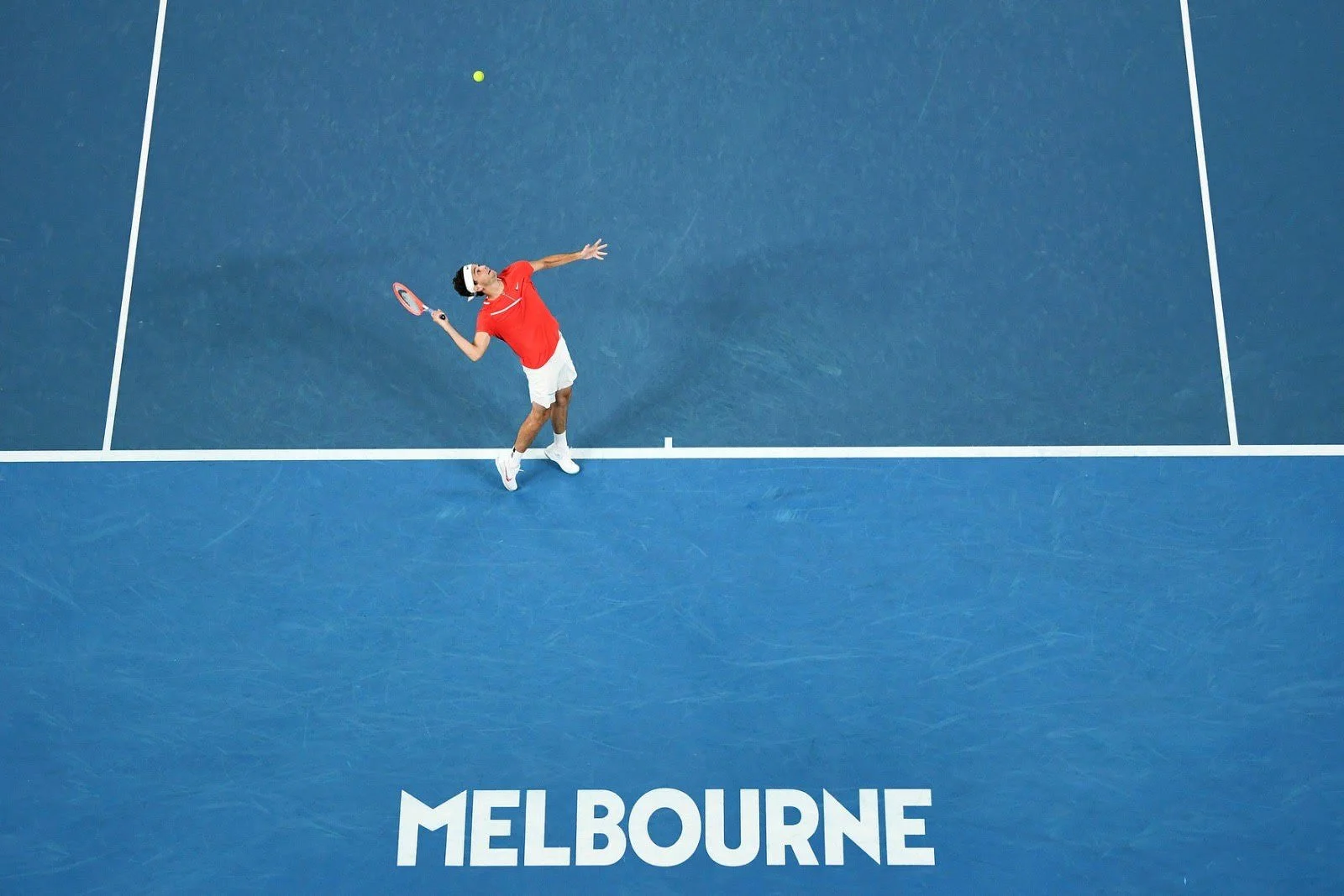 A tennis player in a red shirt and white shorts preparing to hit a yellow tennis ball on a blue tennis court in Melbourne.