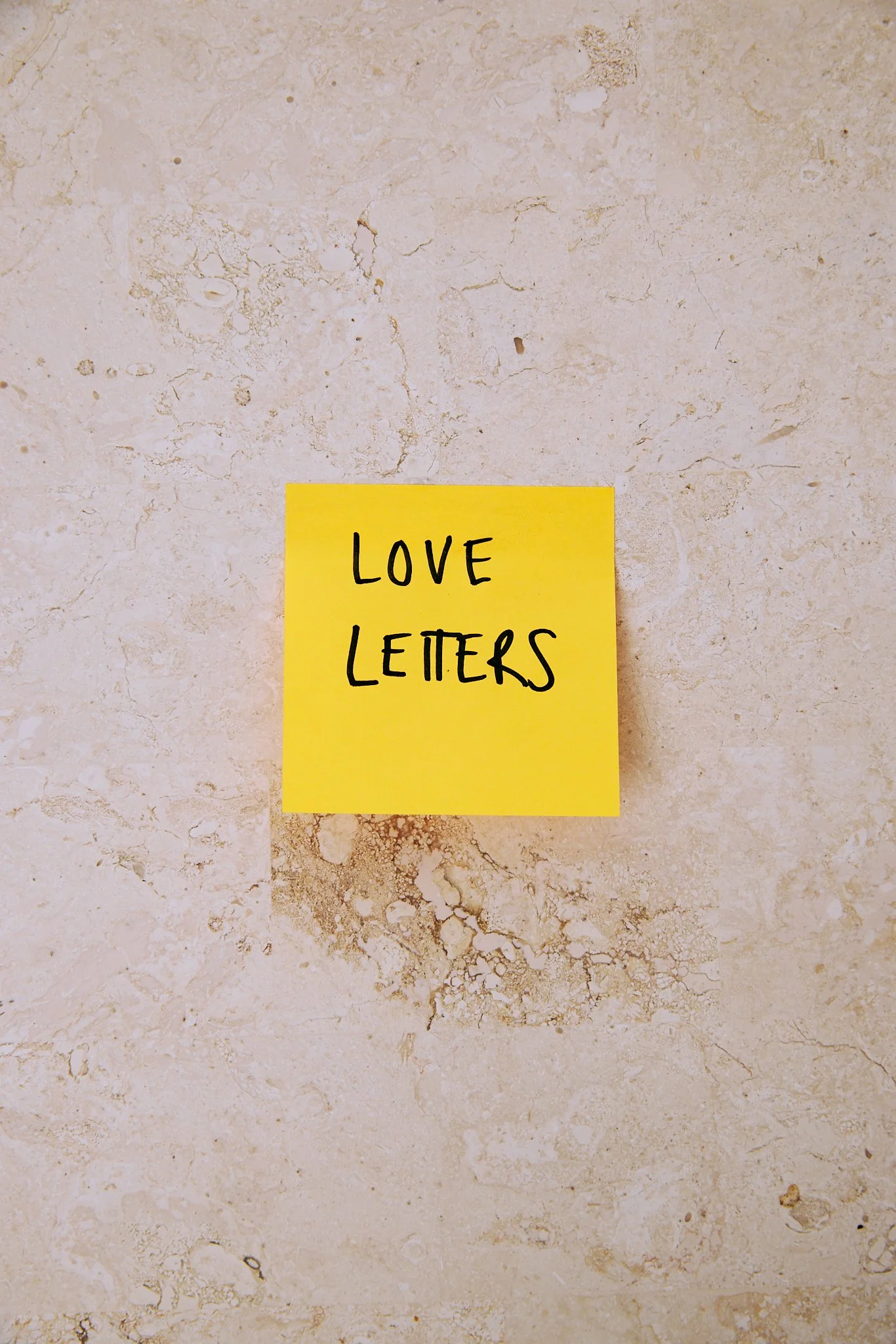 A yellow sticky note with handwritten black text that says 'Love Letters' on a beige textured surface.