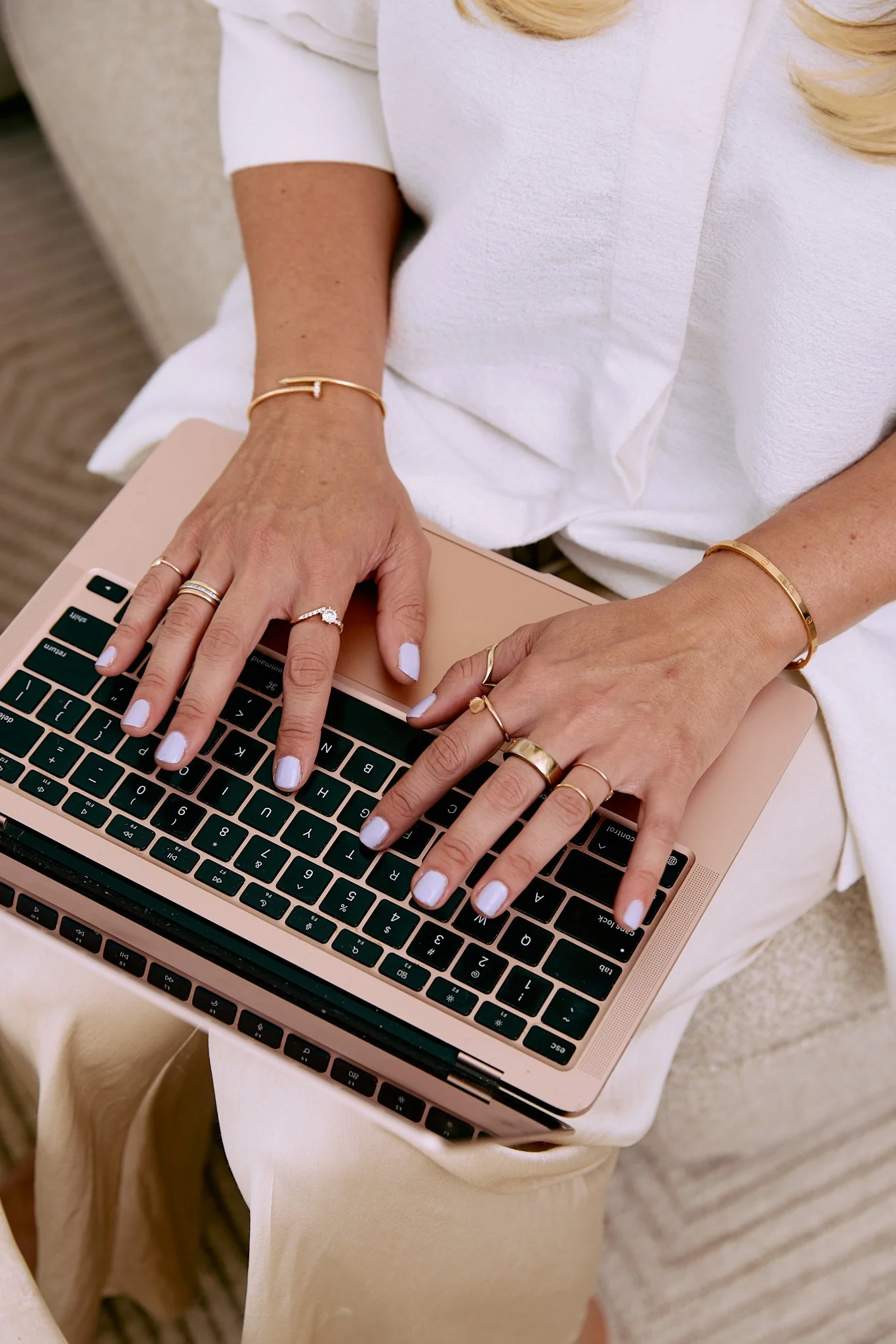 Close-up of a woman’s hands with multiple gold rings and bracelets, typing on a rose gold laptop keyboard, seated on a beige chair.