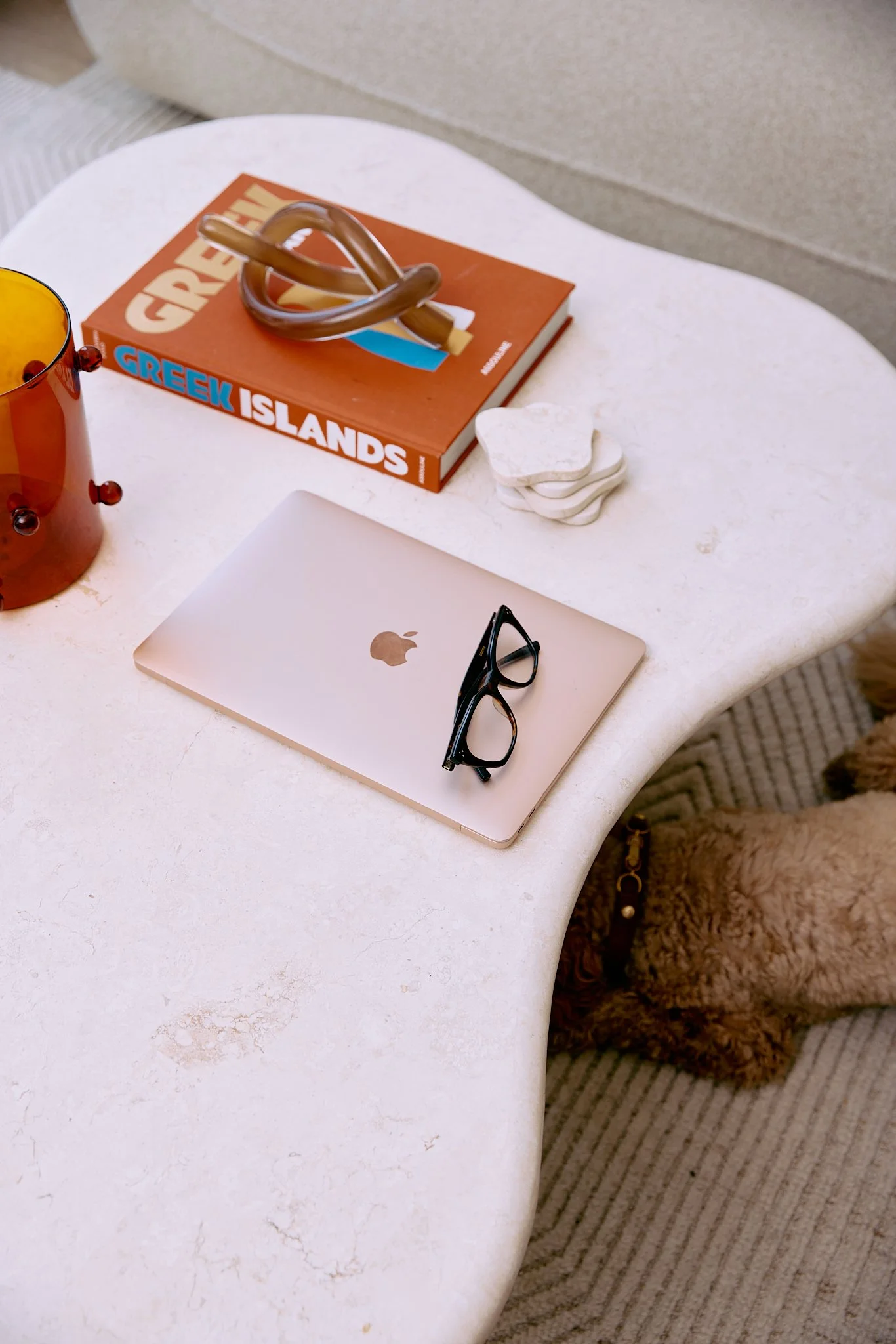 A white, curved table with a closed MacBook, black glasses on top, a book titled 'Greek Islands', white stones, a brown beverage in a glass, and a brown furry dog under the table.