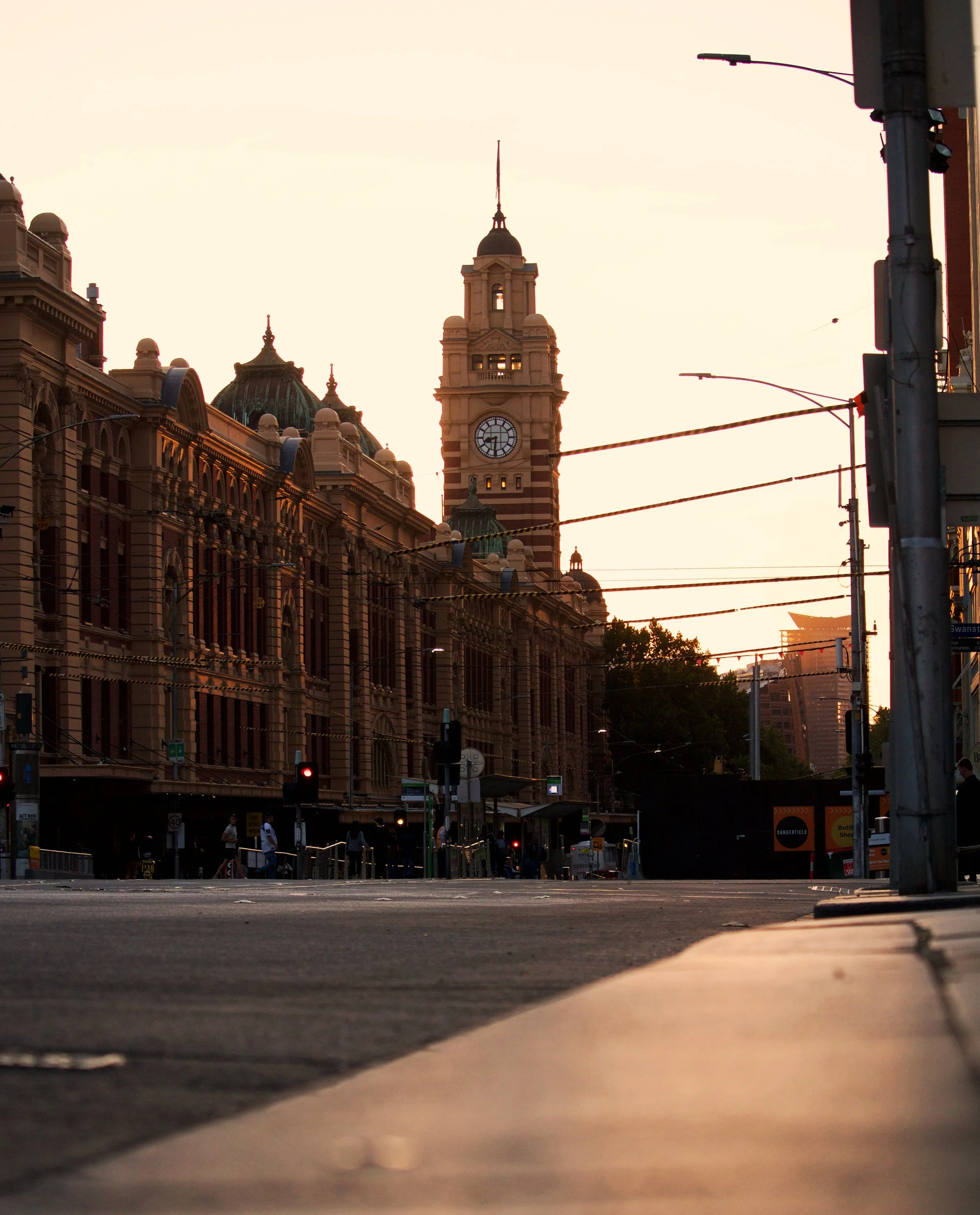 City street during sunset with historic building and clock tower in the background, streetlights, tram wires, and a few pedestrians